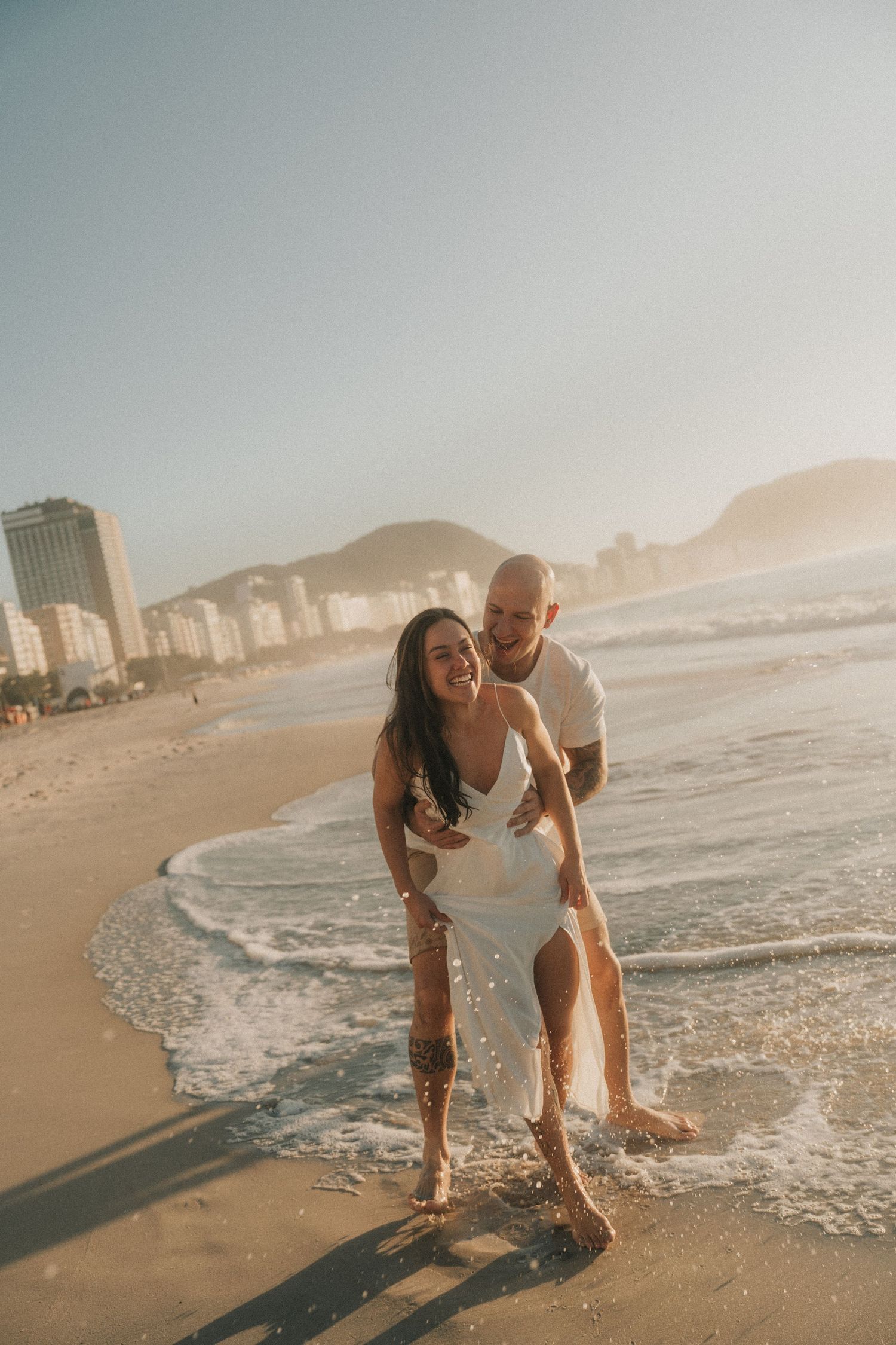A couple shares romantic moments on a scenic beach with city buildings and mountains in the background during golden hour.