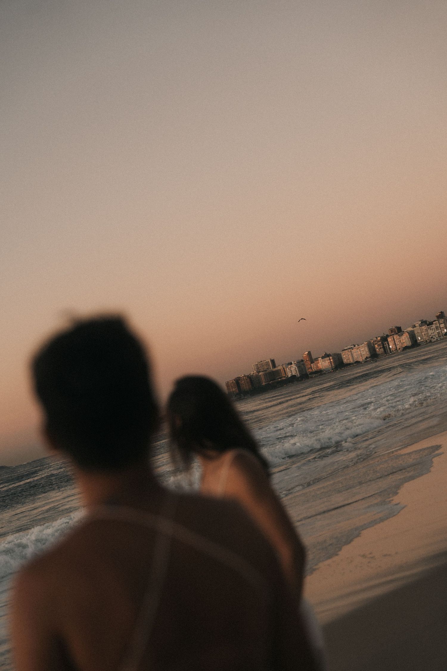 Two silhouetted figures watch the sunset on a beach with city buildings visible along the coastline in soft golden light.