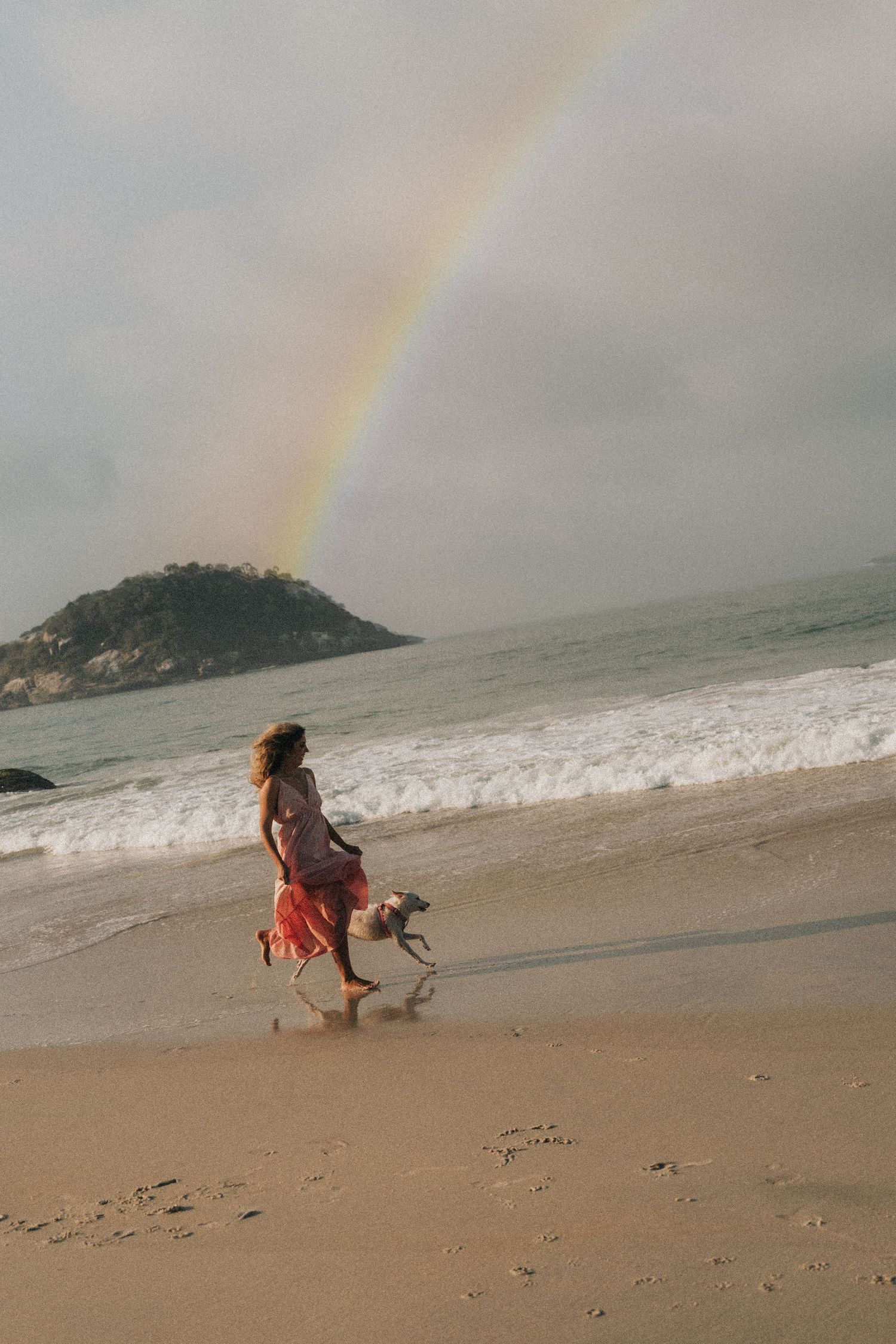 Person in flowing red dress walks dog on beach with rainbow in cloudy sky.