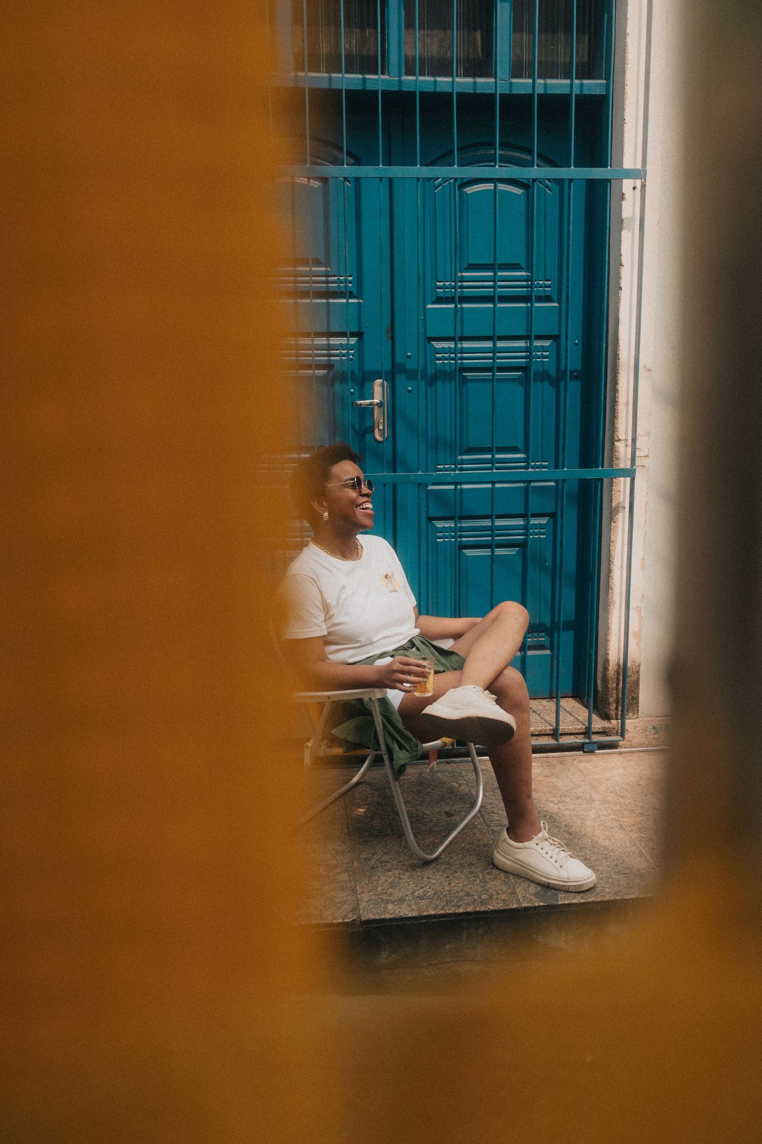Person in white shirt and shorts relaxes in metal folding chair outside turquoise doors in atmospheric lighting.