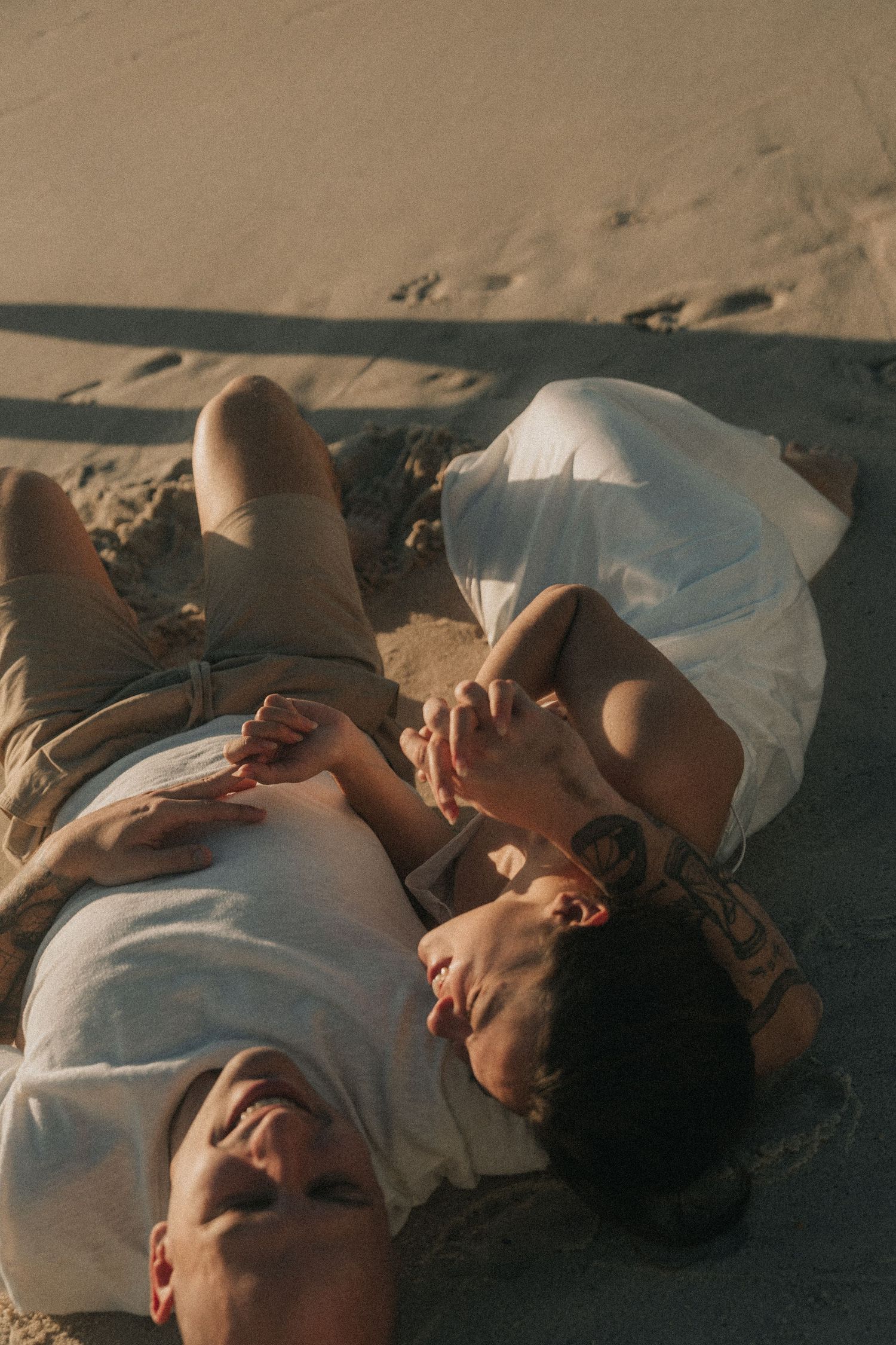 Three people in white clothing lying together on sand at sunset creating intimate artistic composition.