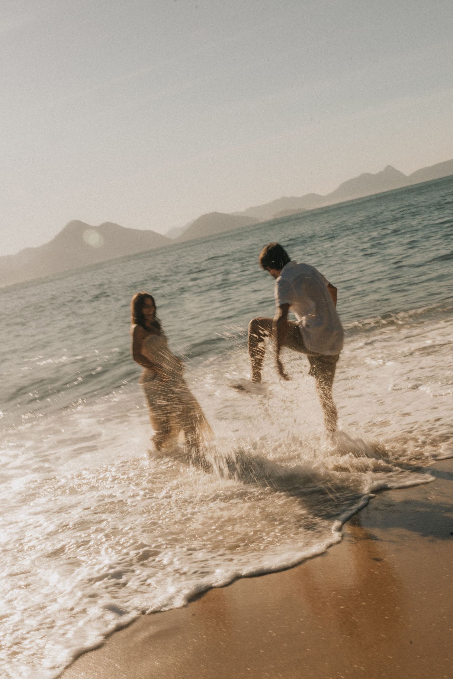 A sequence of playful moments as two people splash and kick in the ocean waves at a beach with mountains in the background.