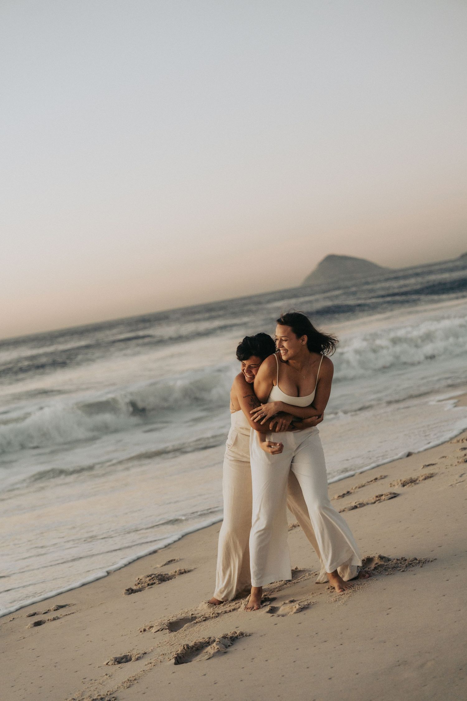 A couple in white outfits embraces on a serene beach at sunset with ocean waves crashing in the background.