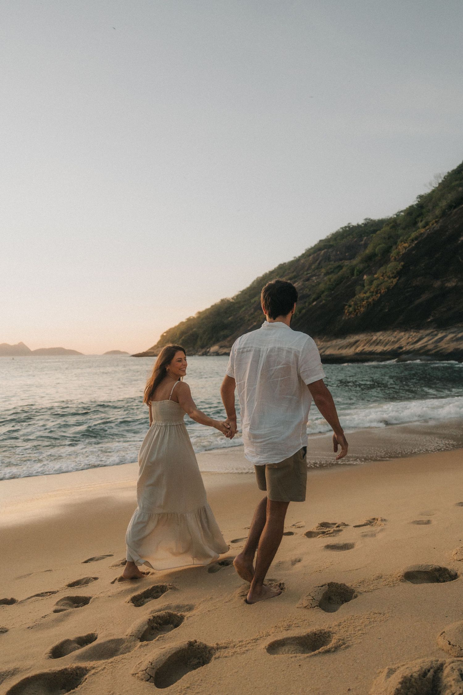 A couple walks hand in hand along a beautiful tropical beach at sunset with mountains and coastline in the background.