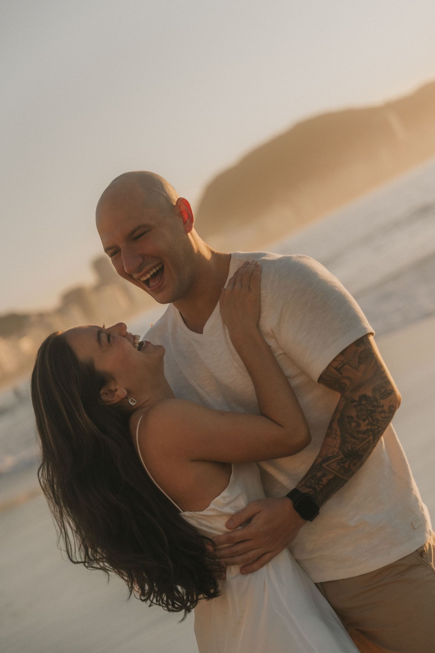 A romantic couple embraces and laughs together during a sunset photoshoot at the beach.