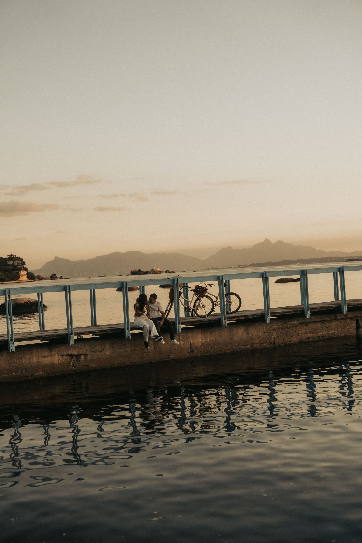 Waterfront dock with silhouettes at sunset against a mountain backdrop and rippling water reflections below.
