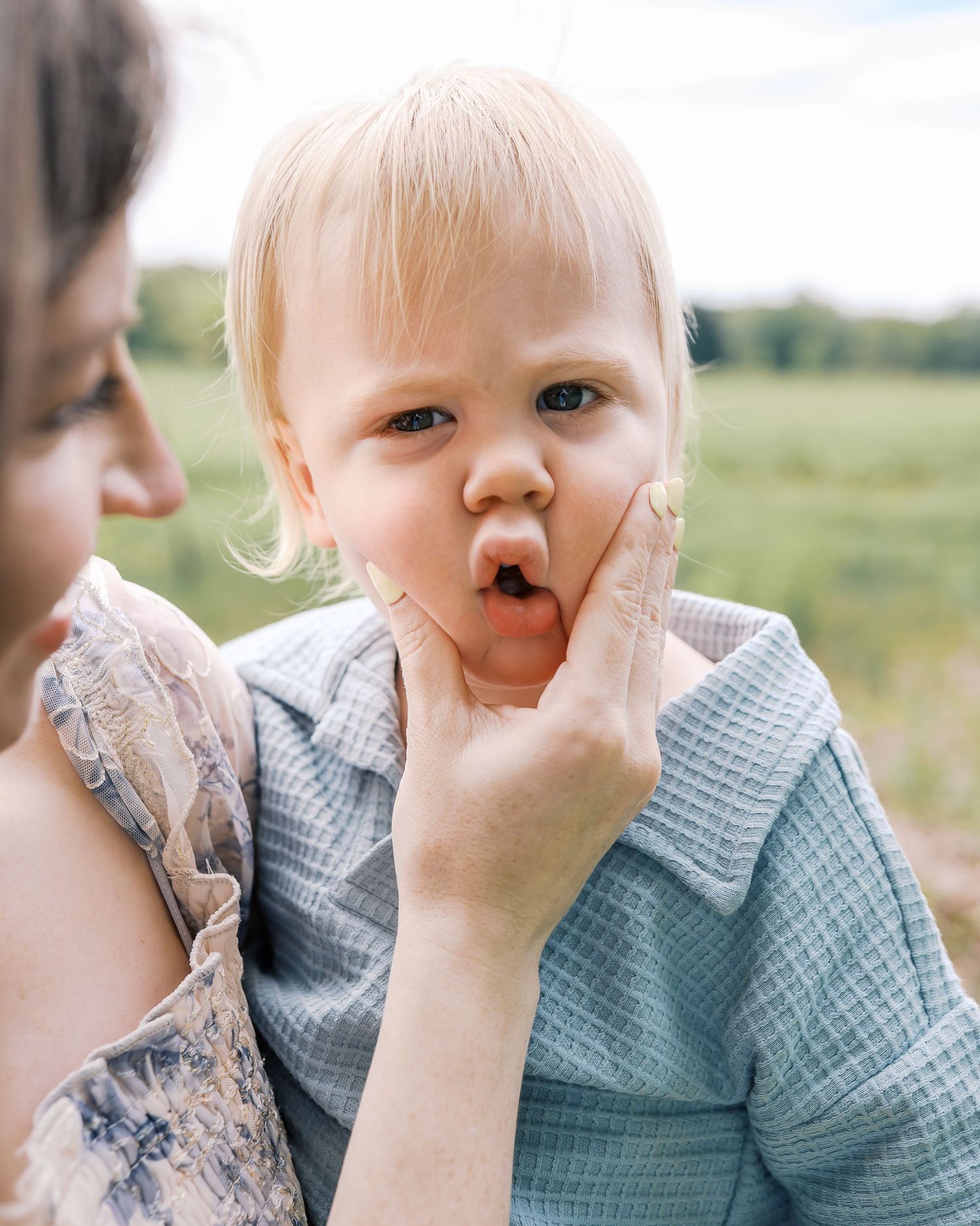 Close-up portrait of a toddler in a checkered outfit touching their face while outdoors.
