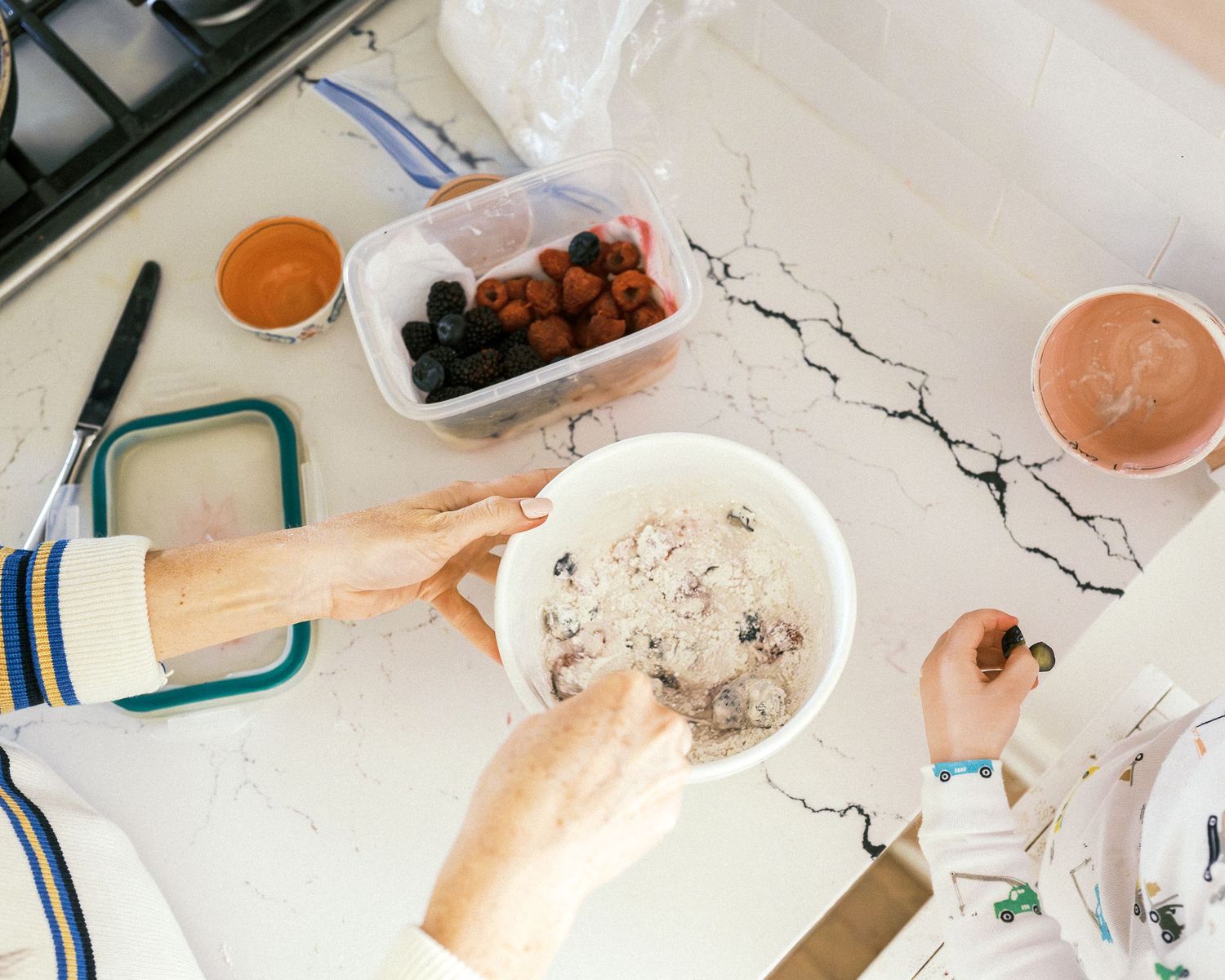 Overhead view of hands preparing food on a white marble countertop with smoothies and ingredients.