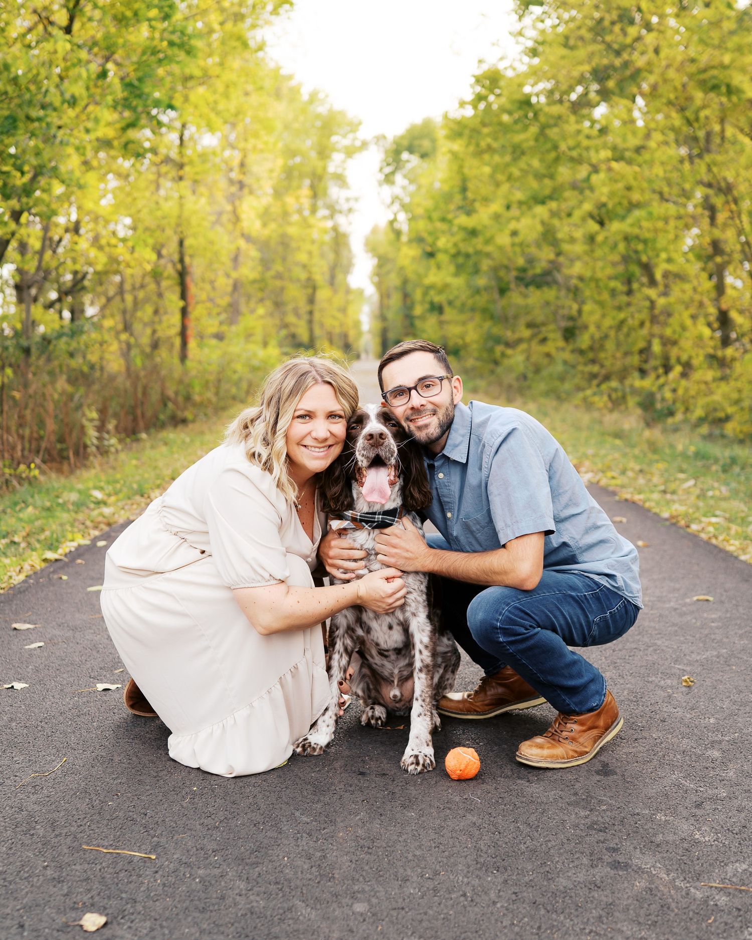 A couple and their spotted dog pose together on a tree-lined path during an autumn photo session.