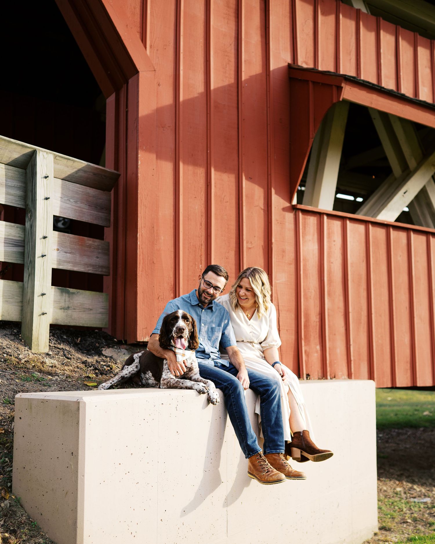 Couple sitting together on a concrete ledge outside a red barn structure in a casual outdoor setting.