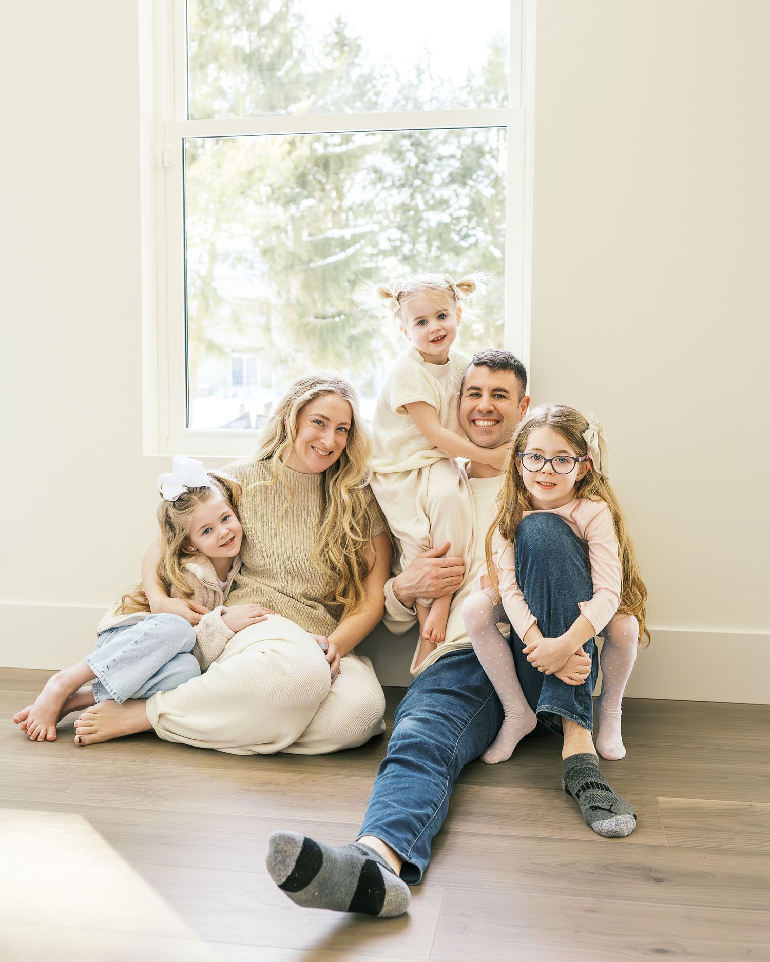 Family members lying playfully on hardwood floor in bright sunlit room with large windows.