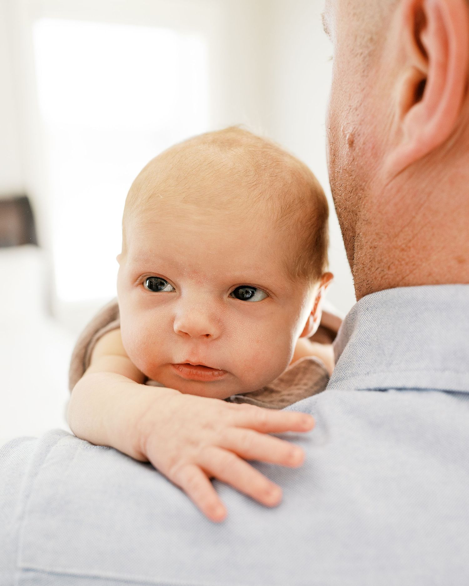 Baby in gray clothing rests on someone's shoulder in natural window light.