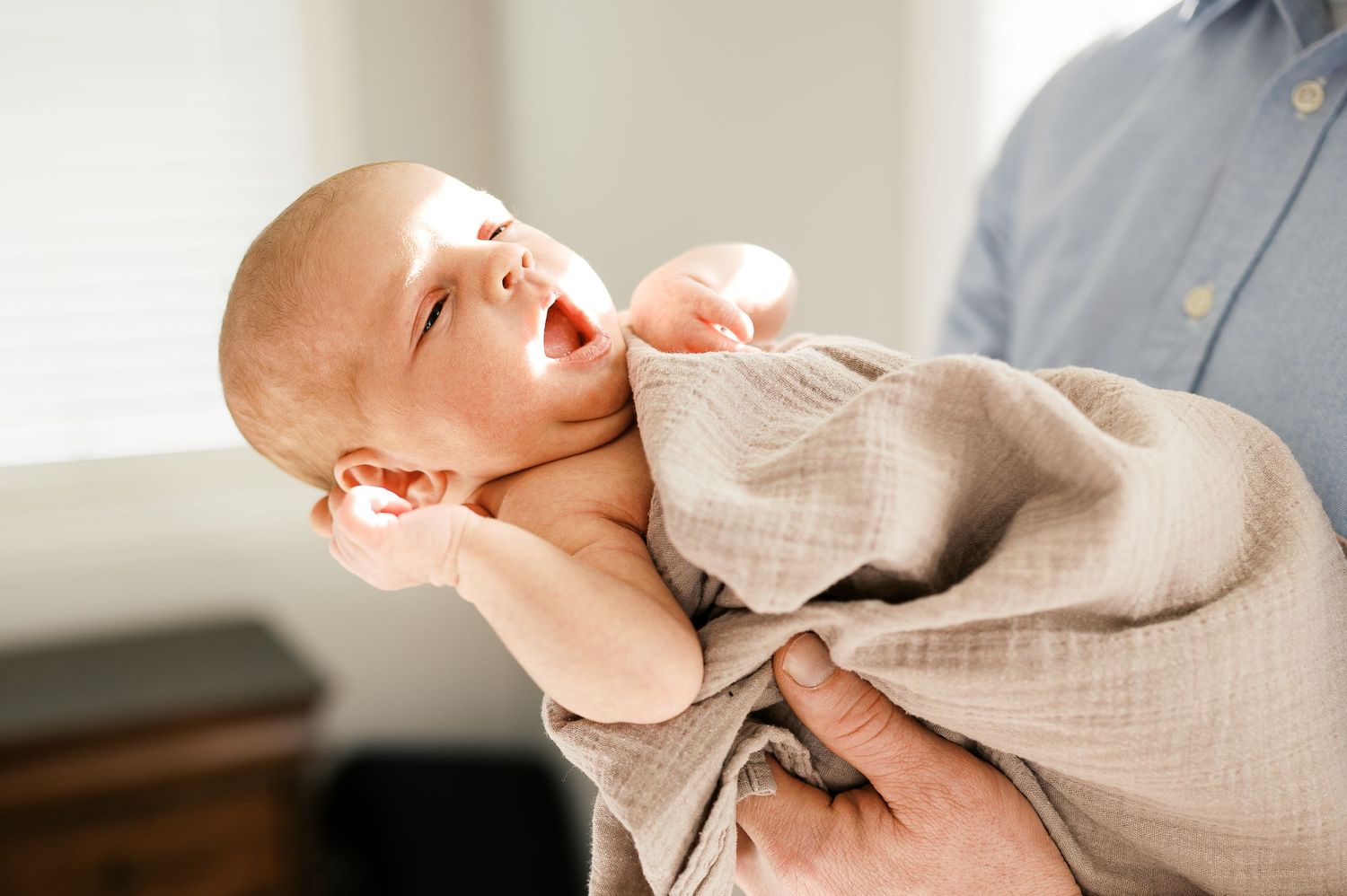 A laughing baby wearing beige clothing is being held in a bright naturally lit room.