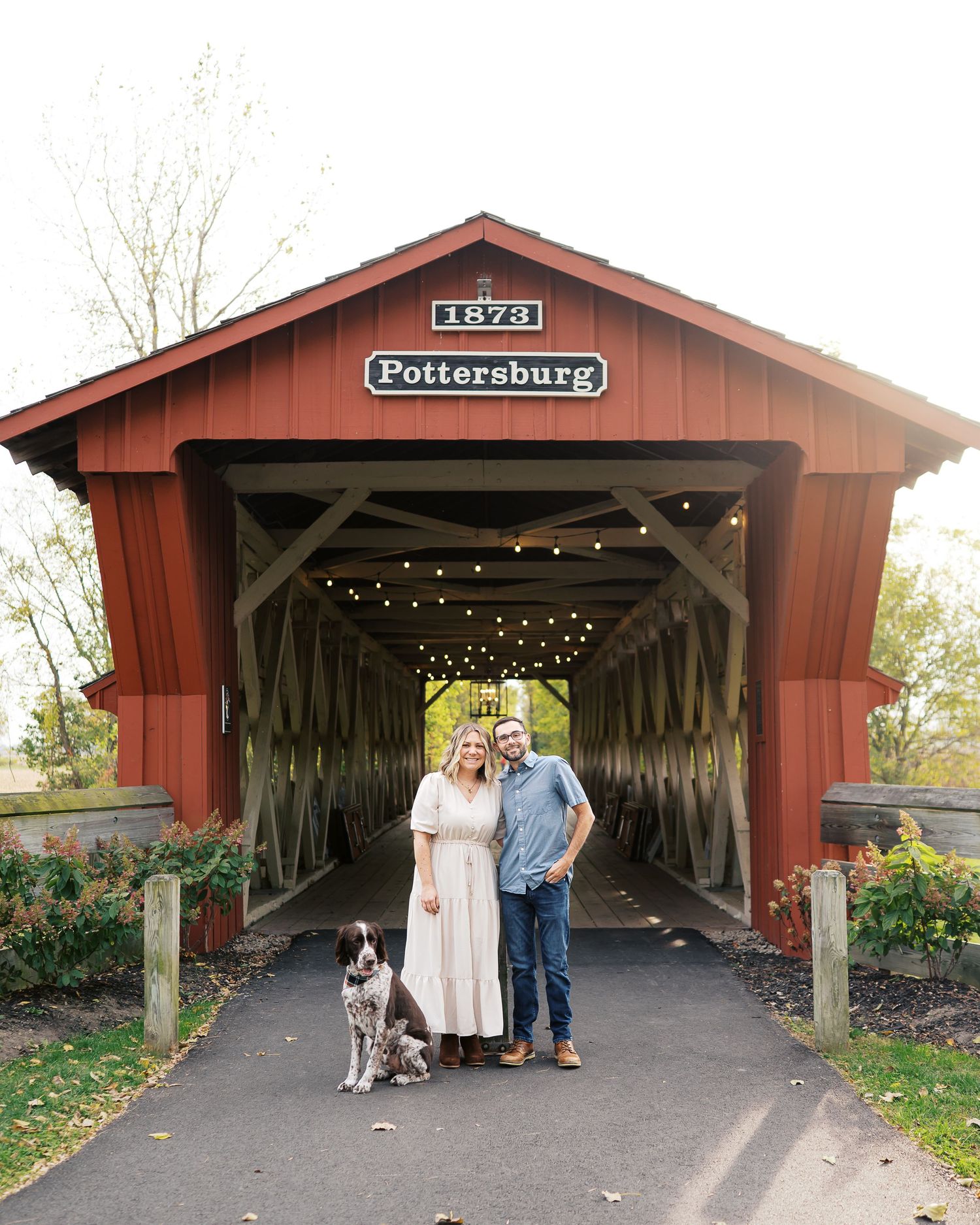 A couple and their dog pose in front of a rustic red covered bridge entrance.