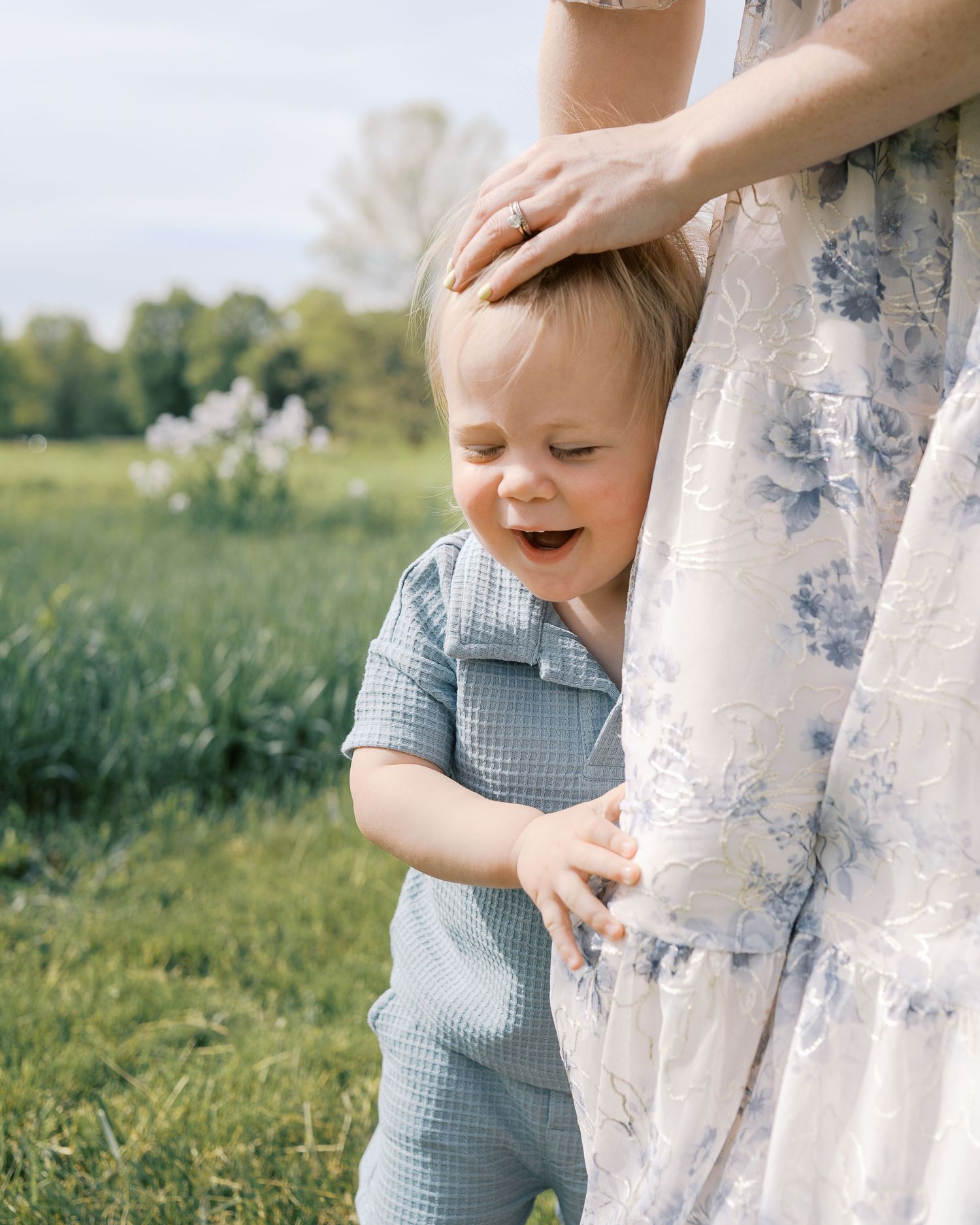 A small child in a blue gingham outfit peeks playfully around a floral dress in a sunny meadow.