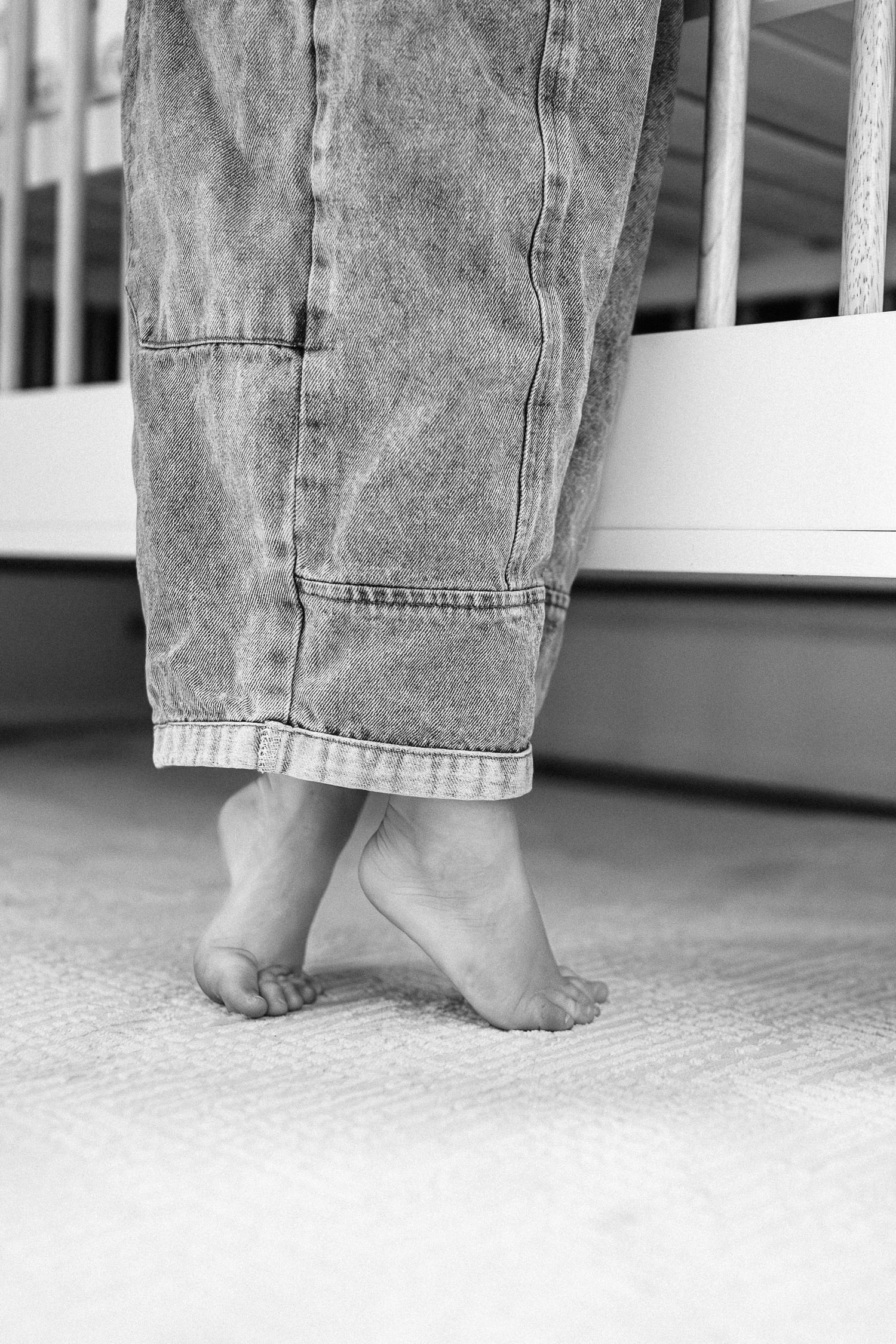 Black and white image of feet in rolled-up denim jeans standing on carpet.