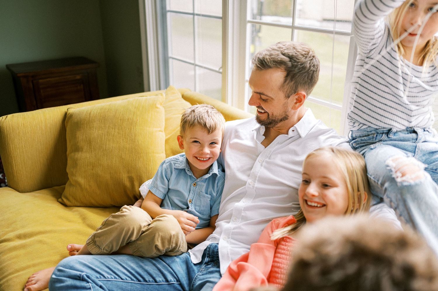 Family members laugh and play together on a yellow couch in a bright room.