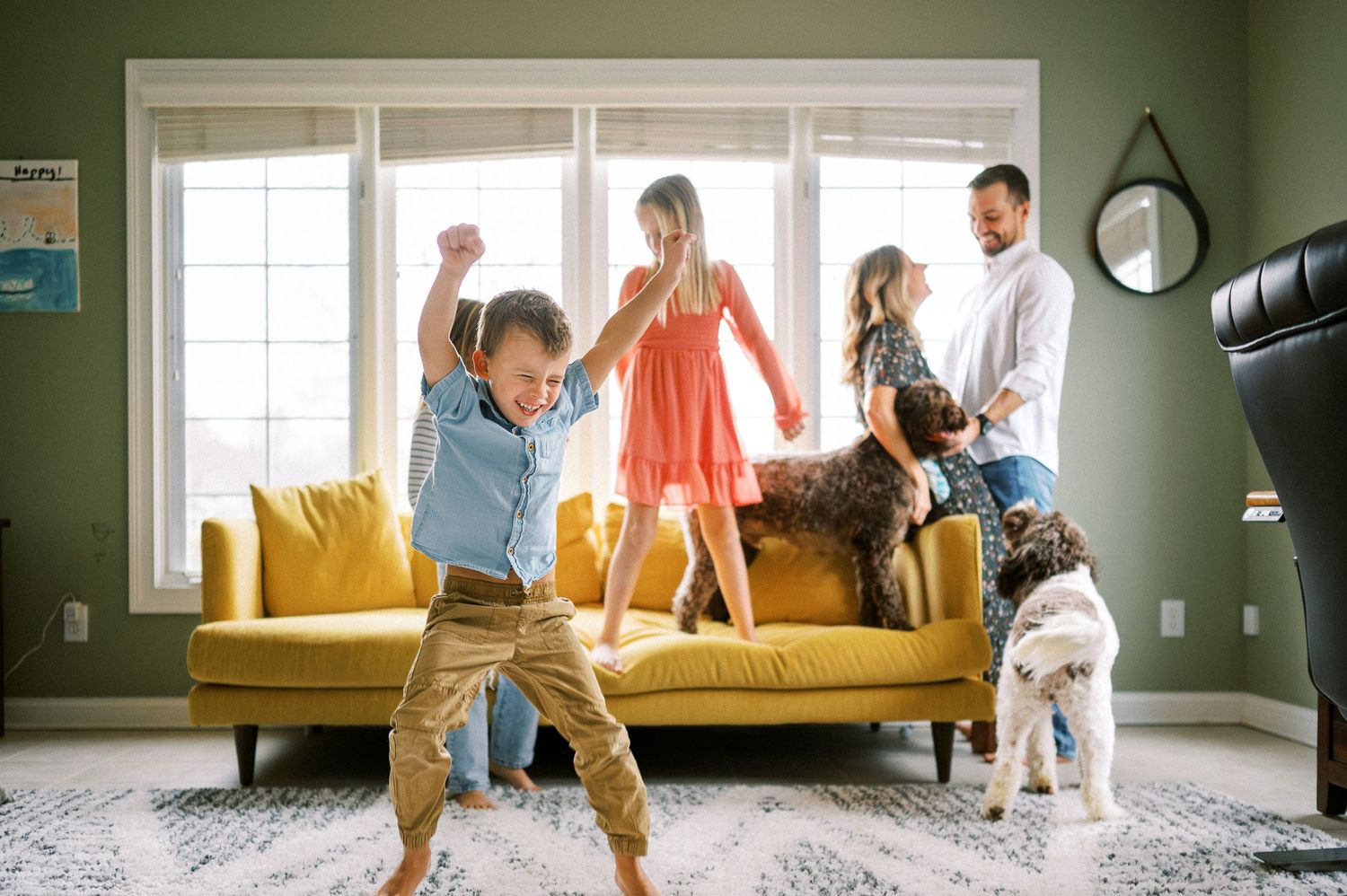 A series of playful family interactions unfold on a yellow couch in front of large windows.