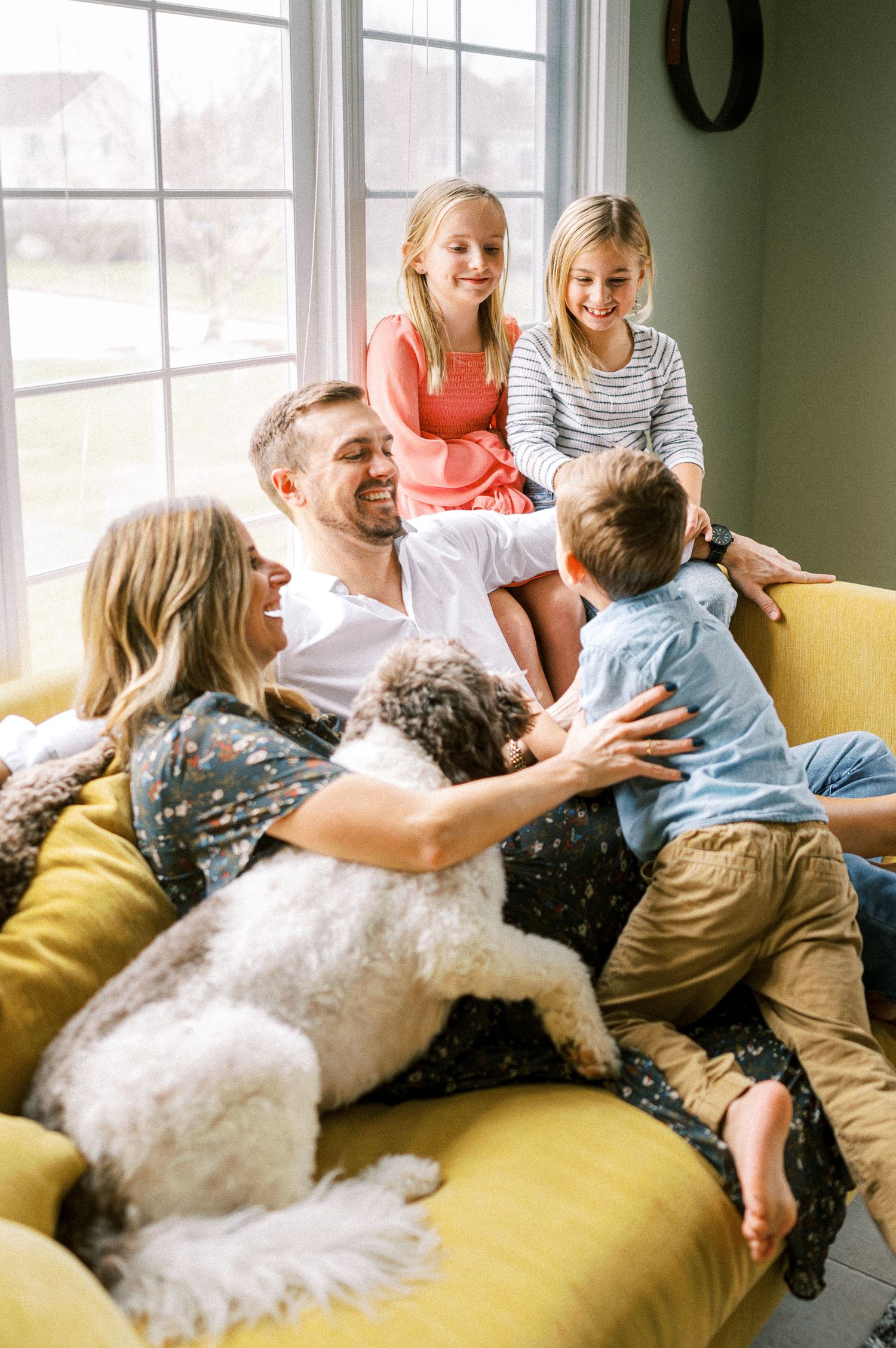 Family members cuddle on a yellow sofa with their fluffy white dog in a sunlit room.