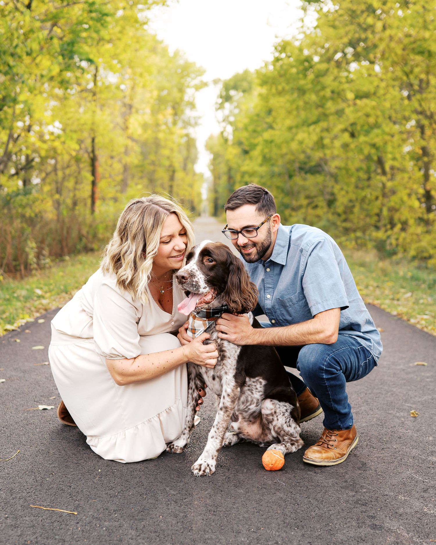 A couple plays with their brown and white spotted dog on a tree-lined path during autumn.