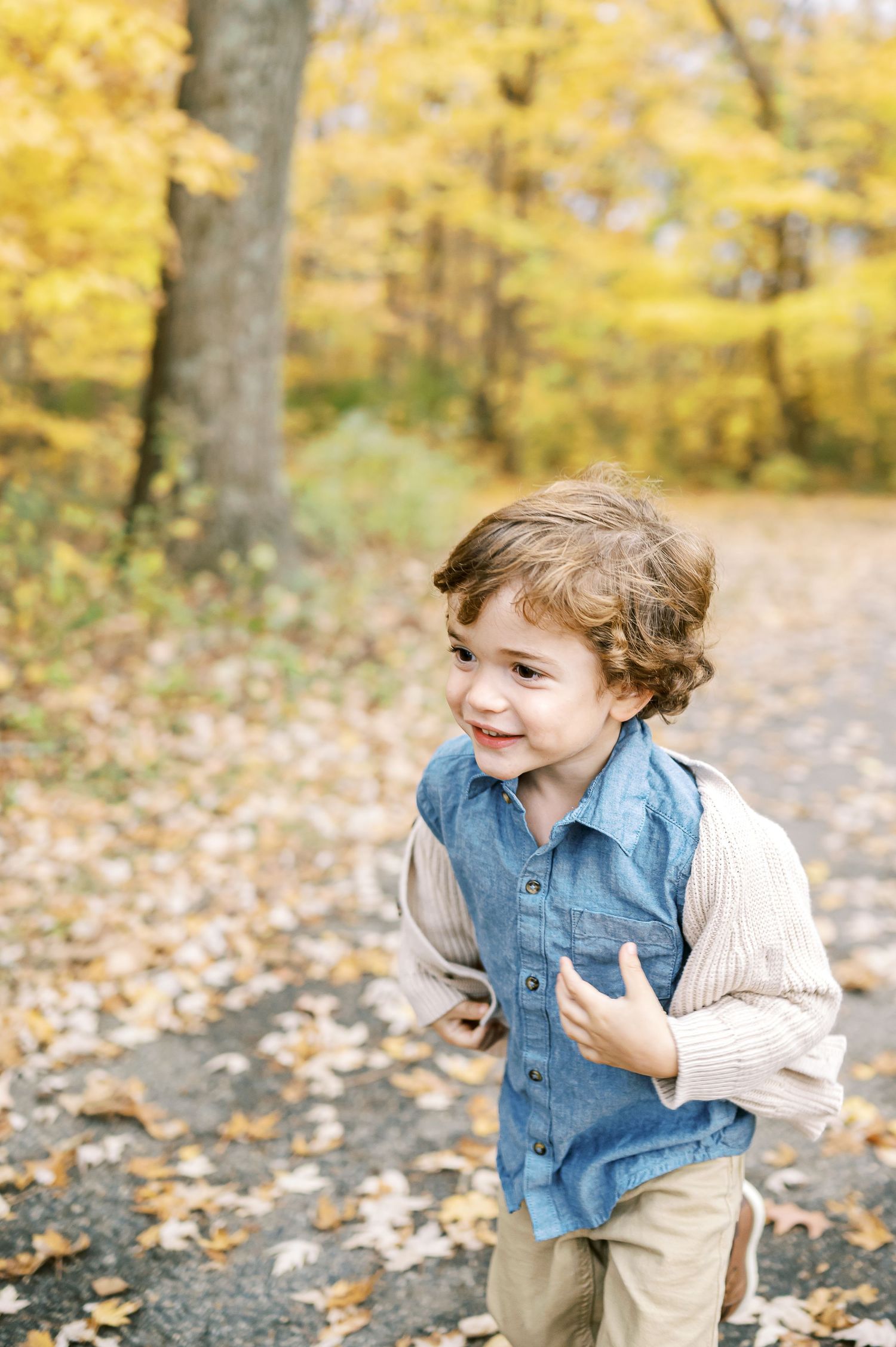 Joyful child wearing denim vest and sweater plays in autumn forest surrounded by golden leaves.