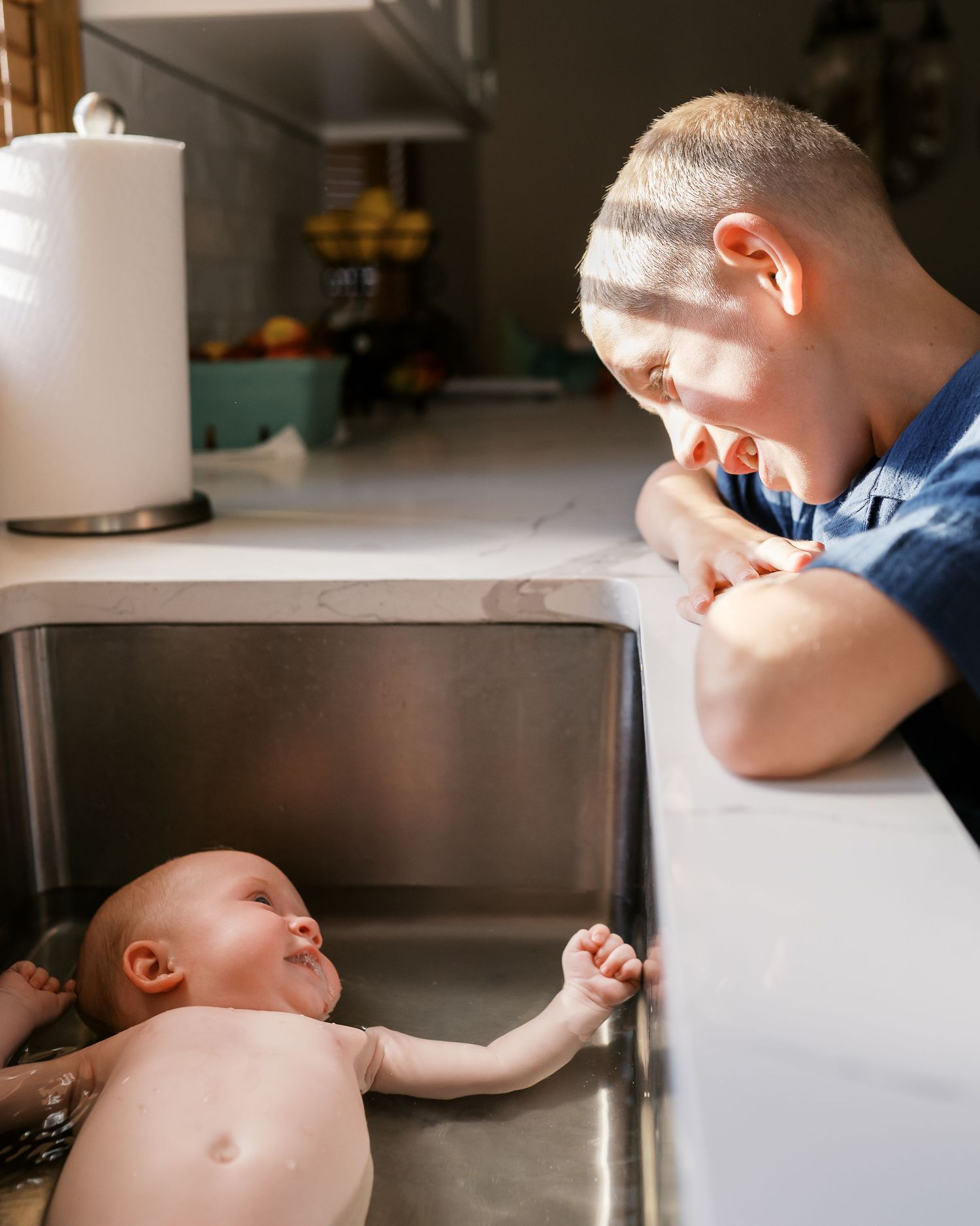 Baby enjoying bath time in stainless steel kitchen sink while sibling watches.