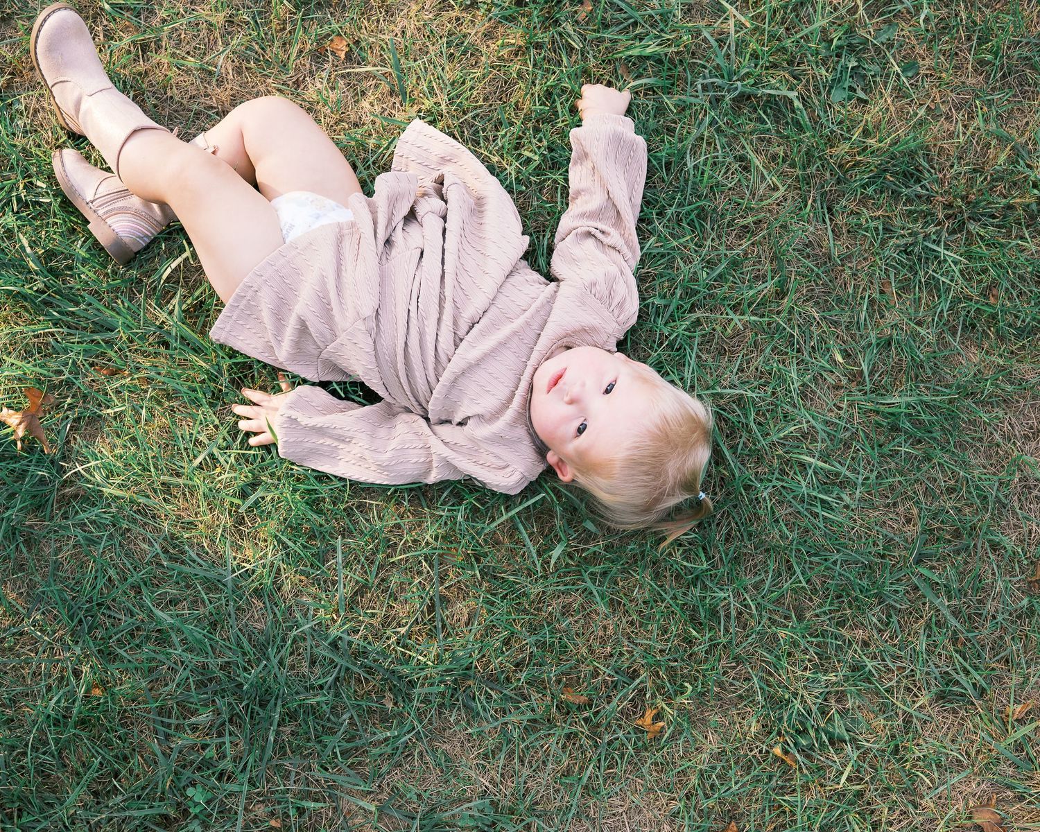 A small figure in light colored clothing lying playfully on green grass during a sunny day.