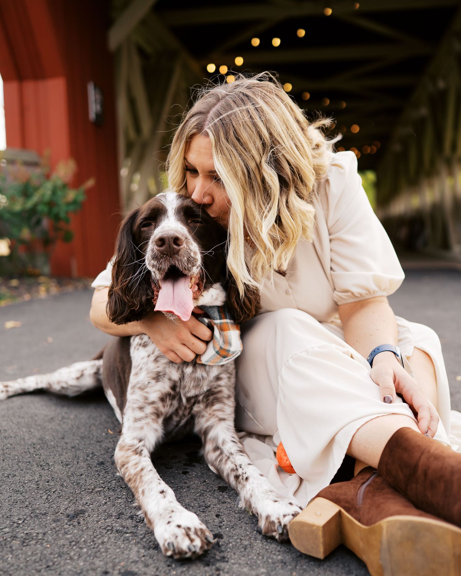 Couple in white clothing cuddling with their spotted dog on ground with bokeh lights in background.