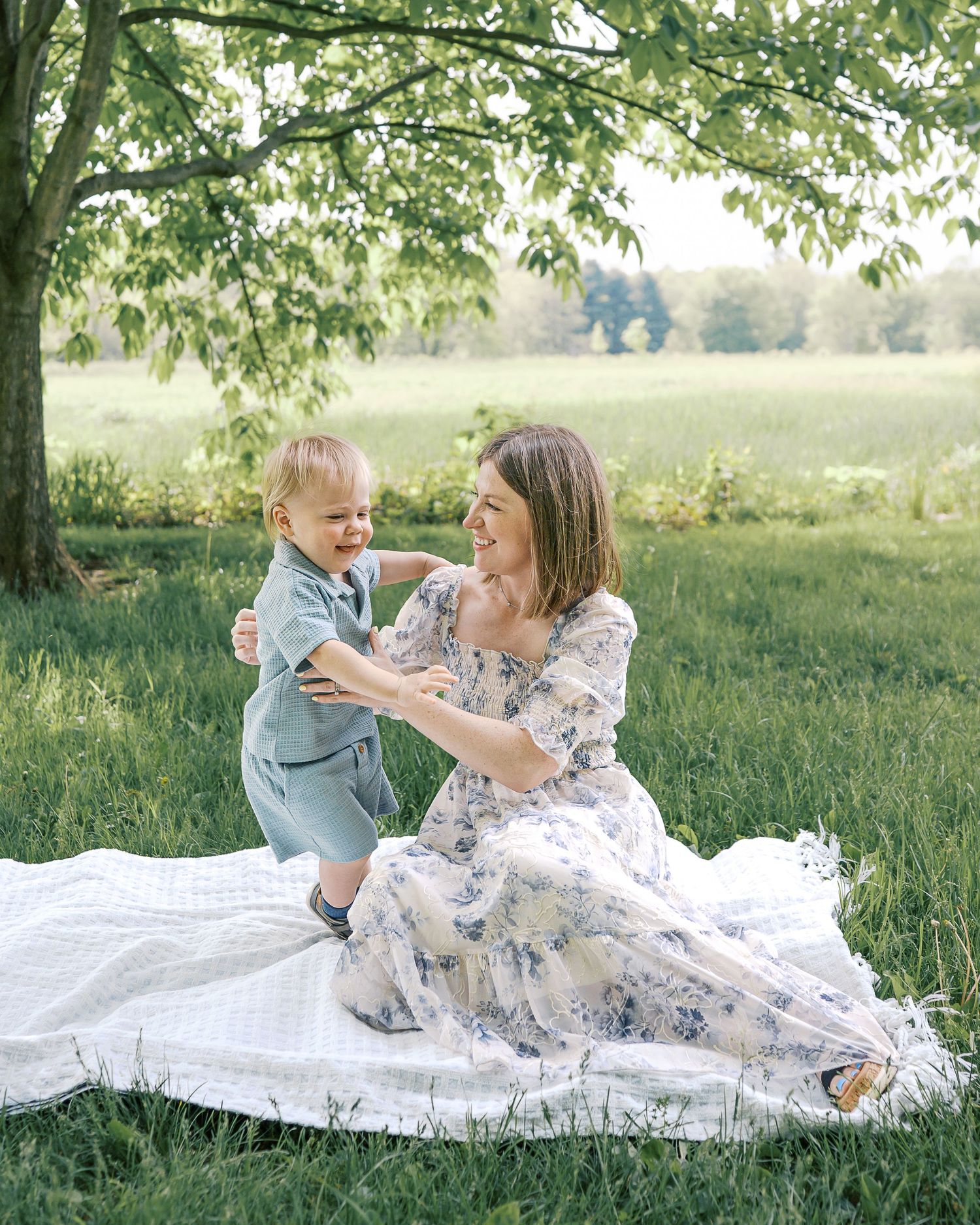A family picnic scene with floral dress and blanket under a green tree.
