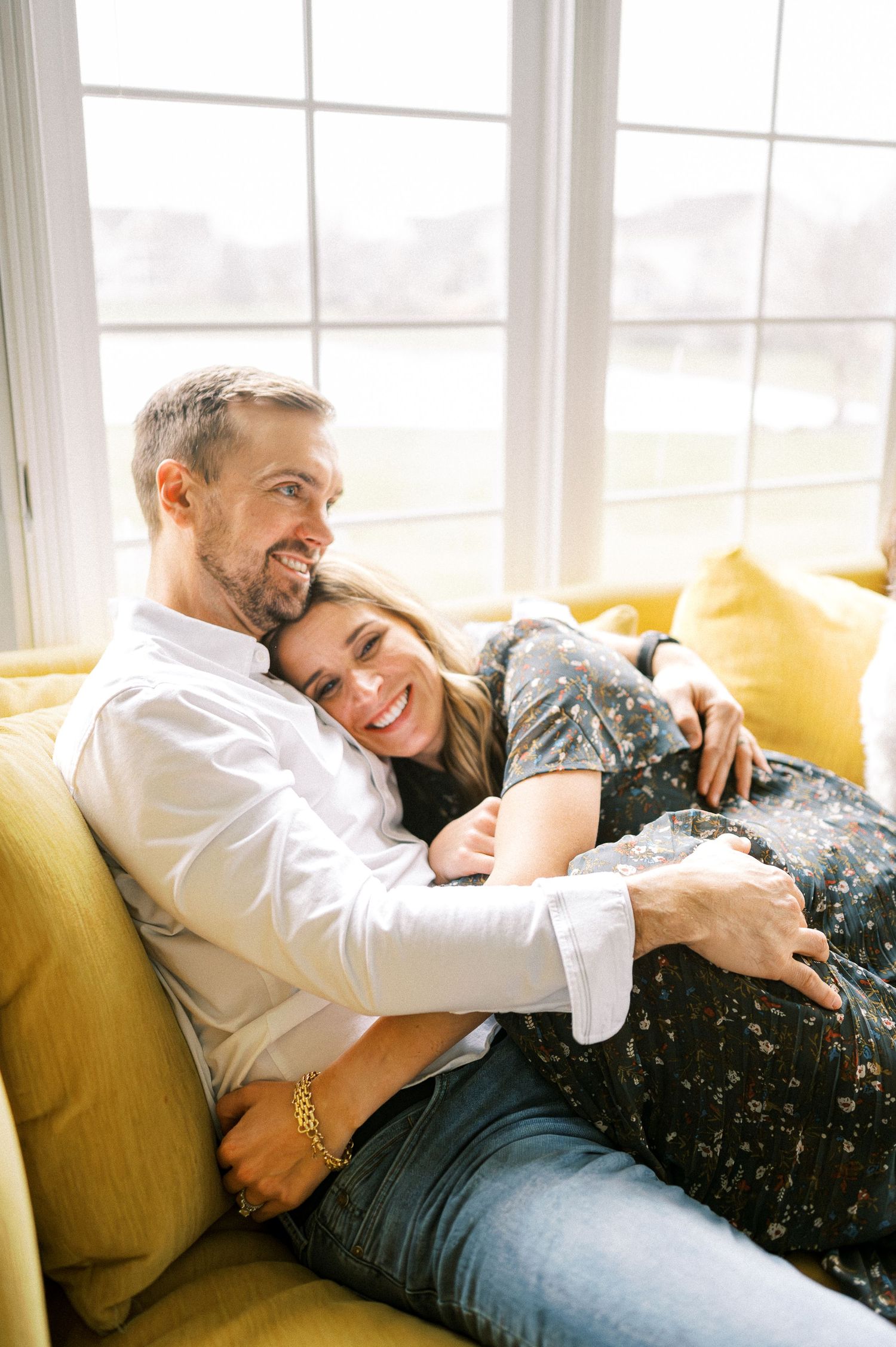 A couple shares a loving embrace while relaxing together on a yellow couch by a bright window.