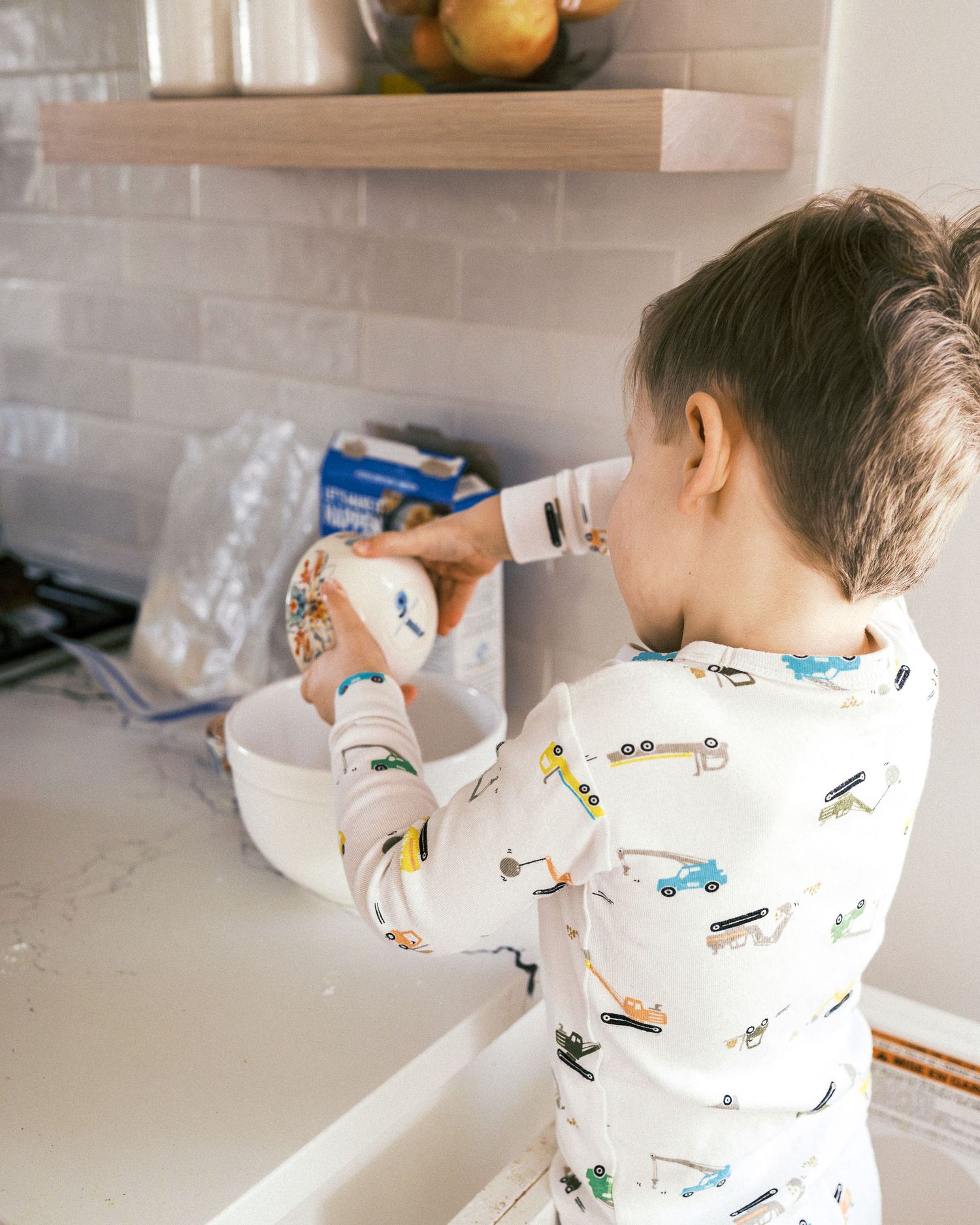 A small child in a patterned white pajama top mixes ingredients in a white bowl on a kitchen counter.