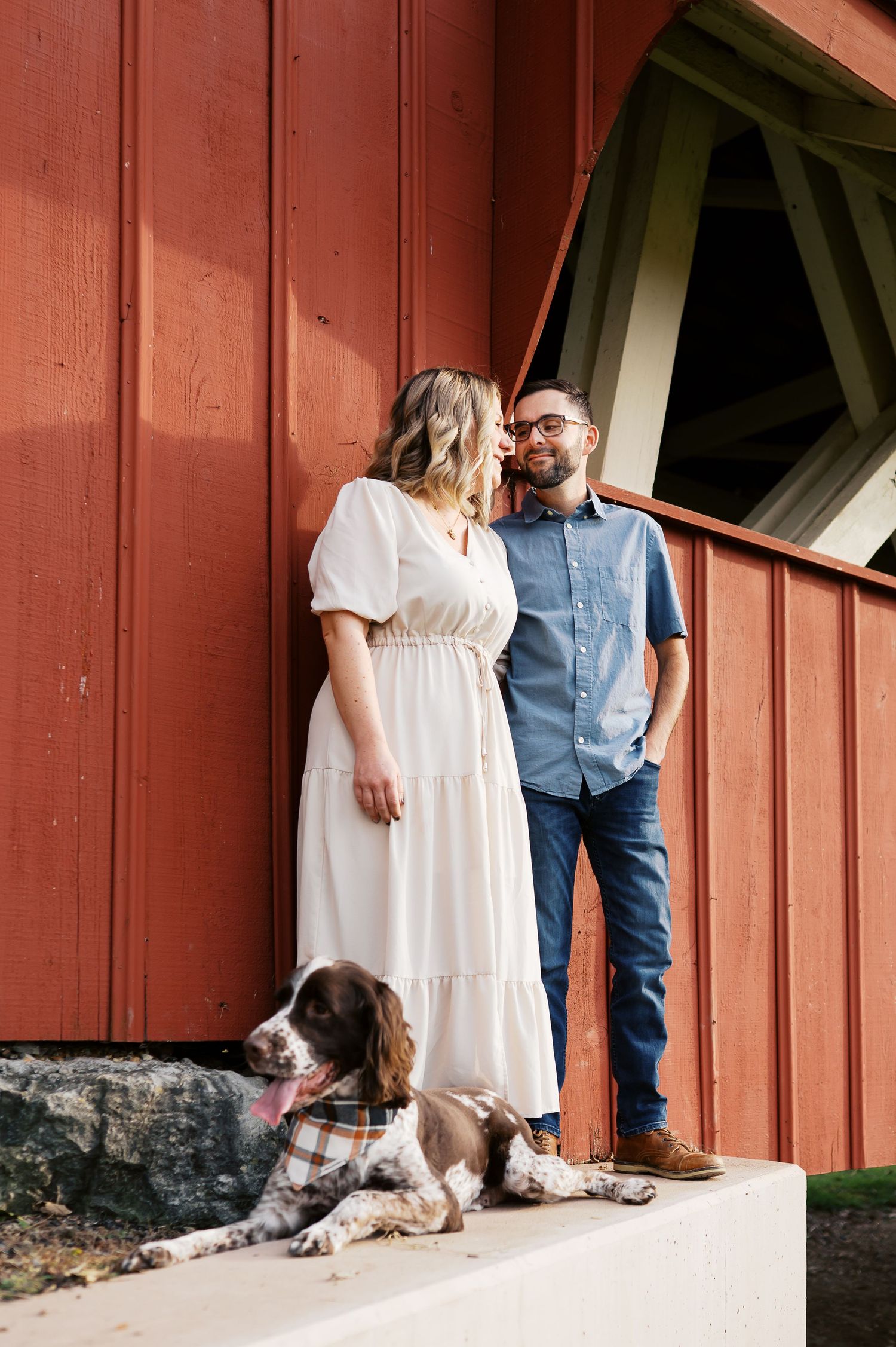 A couple in casual attire stands against a rustic red barn wall with their dog in a series of intimate poses.