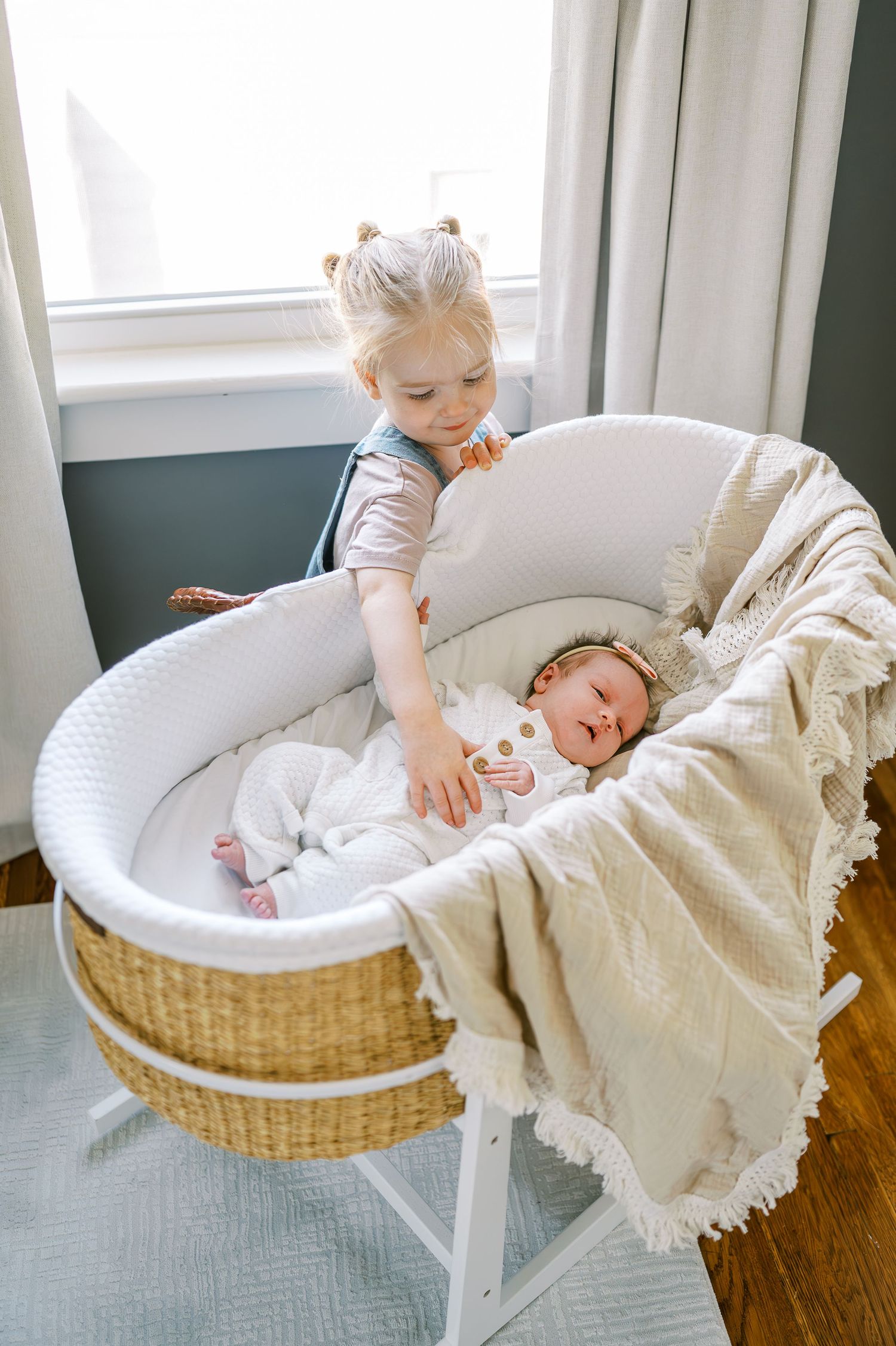 Children near wicker bassinet by window with white curtains.