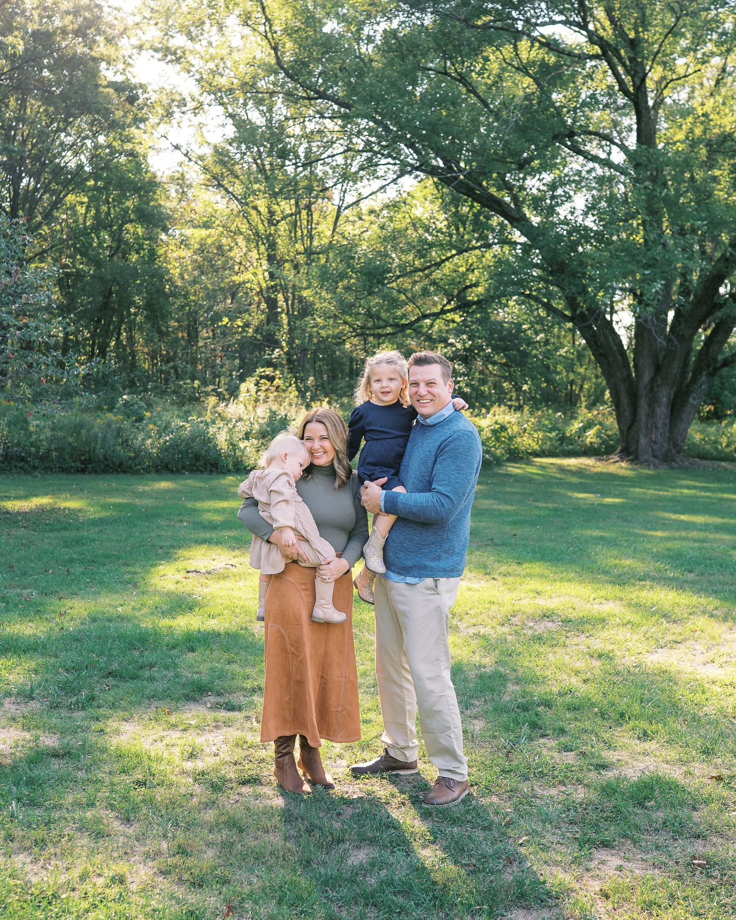 Family poses together in a grassy field with sunlit trees in the background during golden hour.