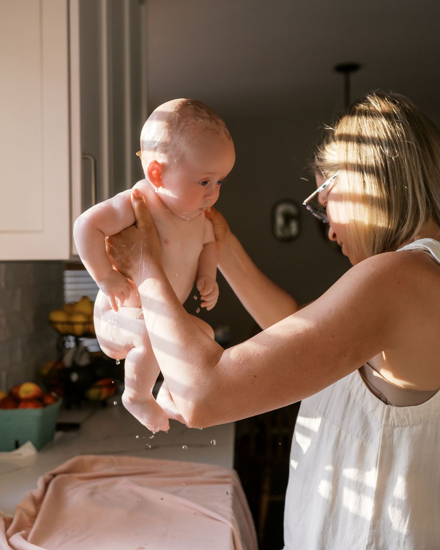 A tender moment in a sunlit kitchen shows a baby being lifted up while wearing only a diaper.