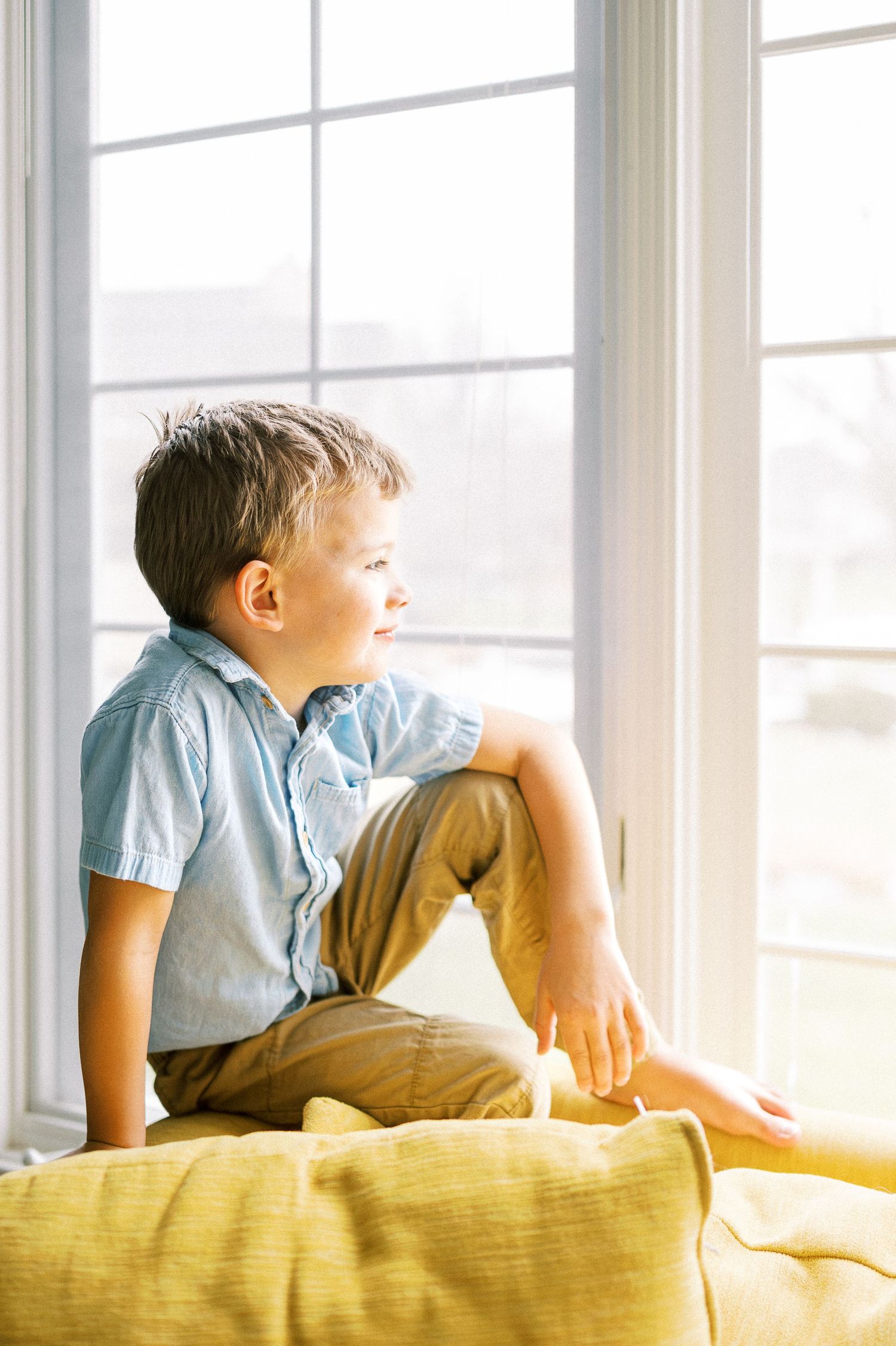 A young child lounges on a yellow couch by a bright window wearing a light blue shirt.