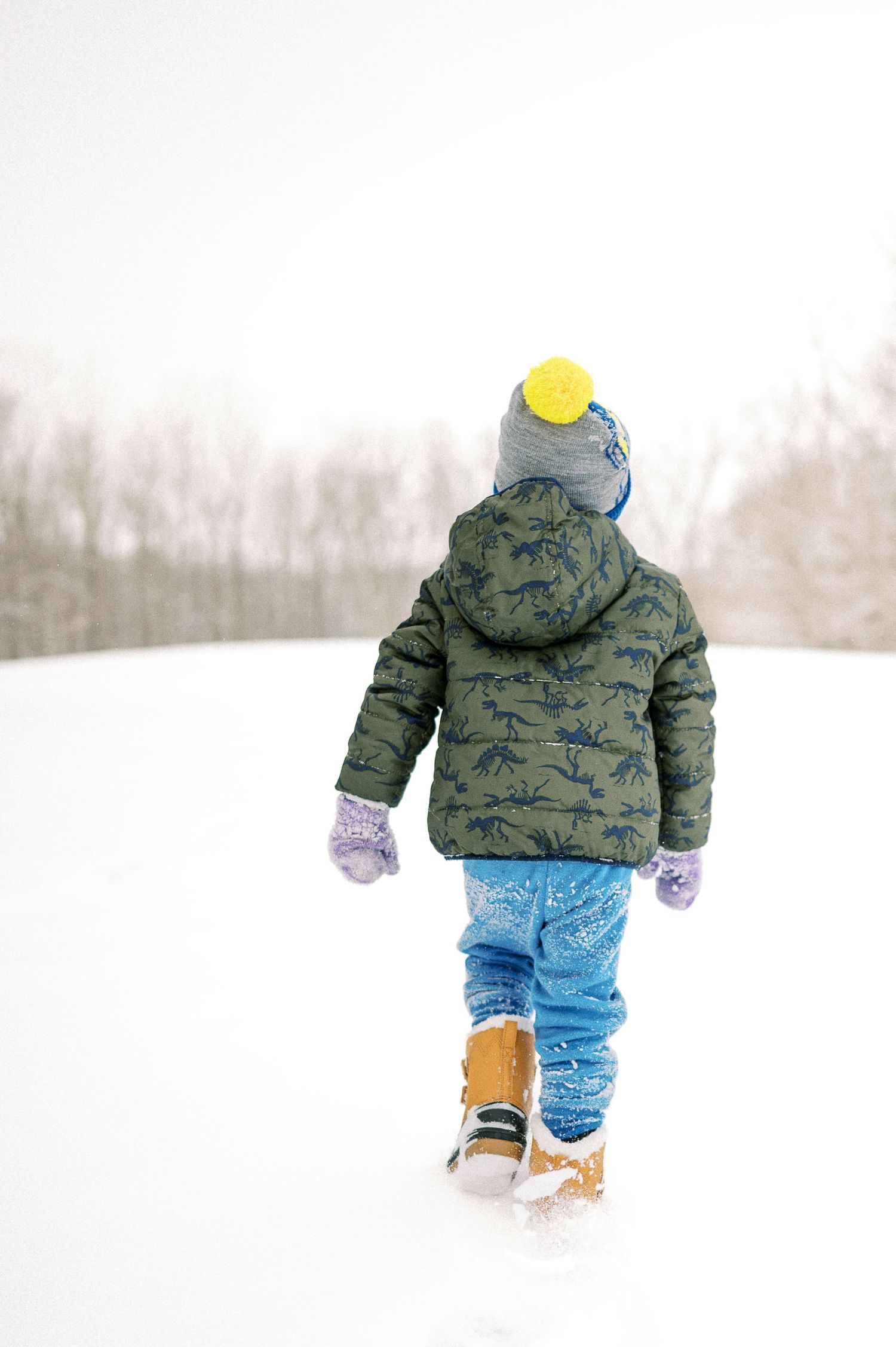 A small figure walks through deep snow wearing a patterned winter coat and yellow hat.