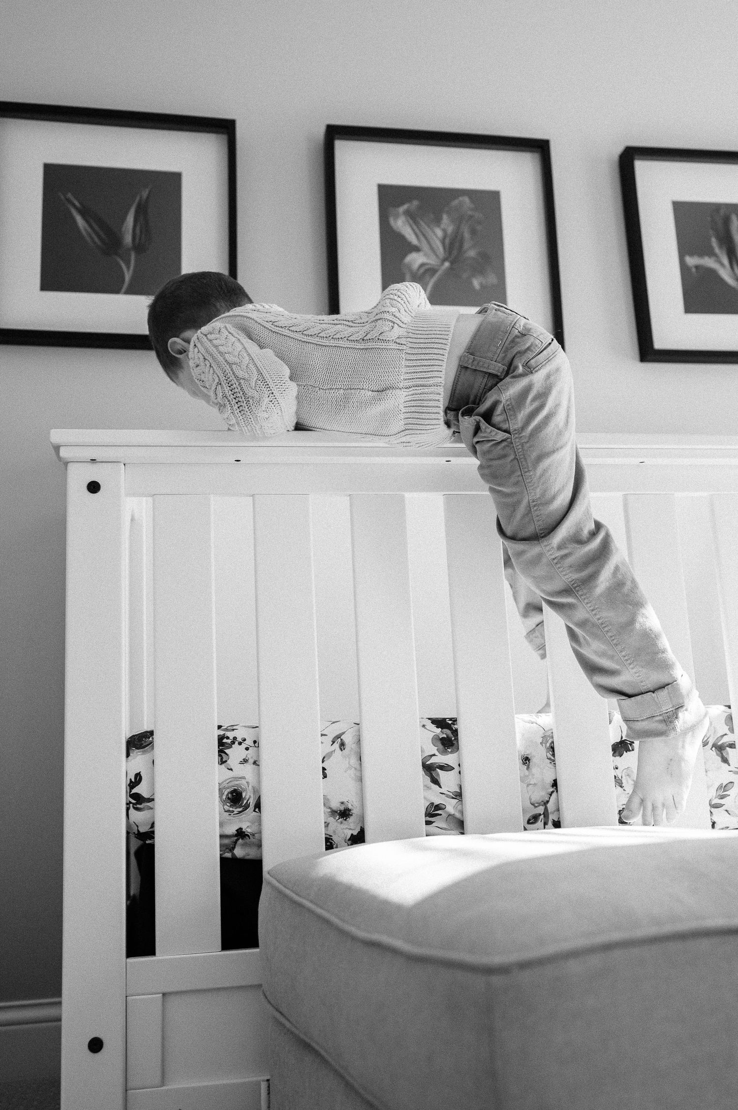 Black and white photo of child climbing on white crib under framed wall art.