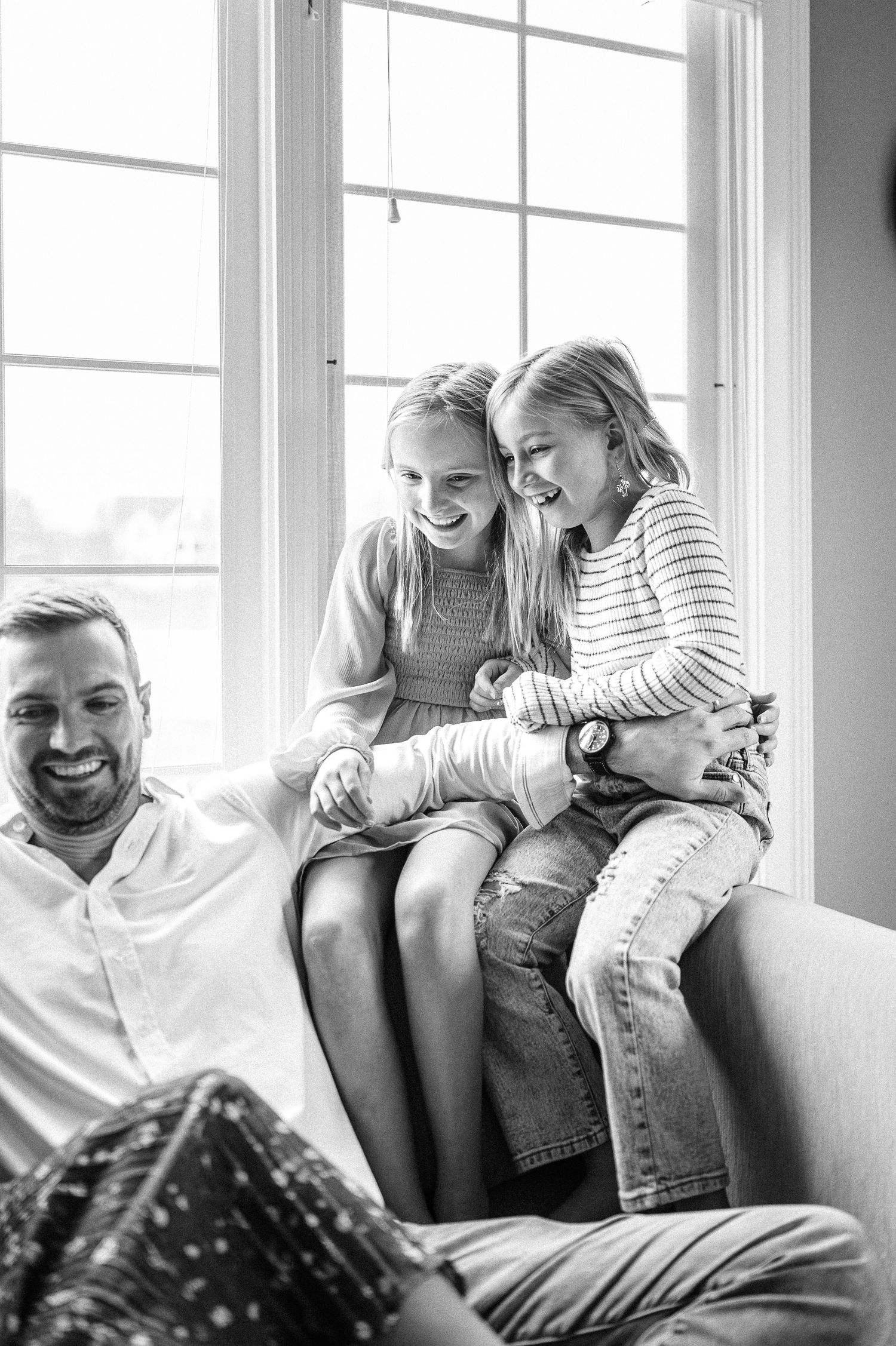 Family members share laughter and smiles while relaxing on a couch near bright windows.