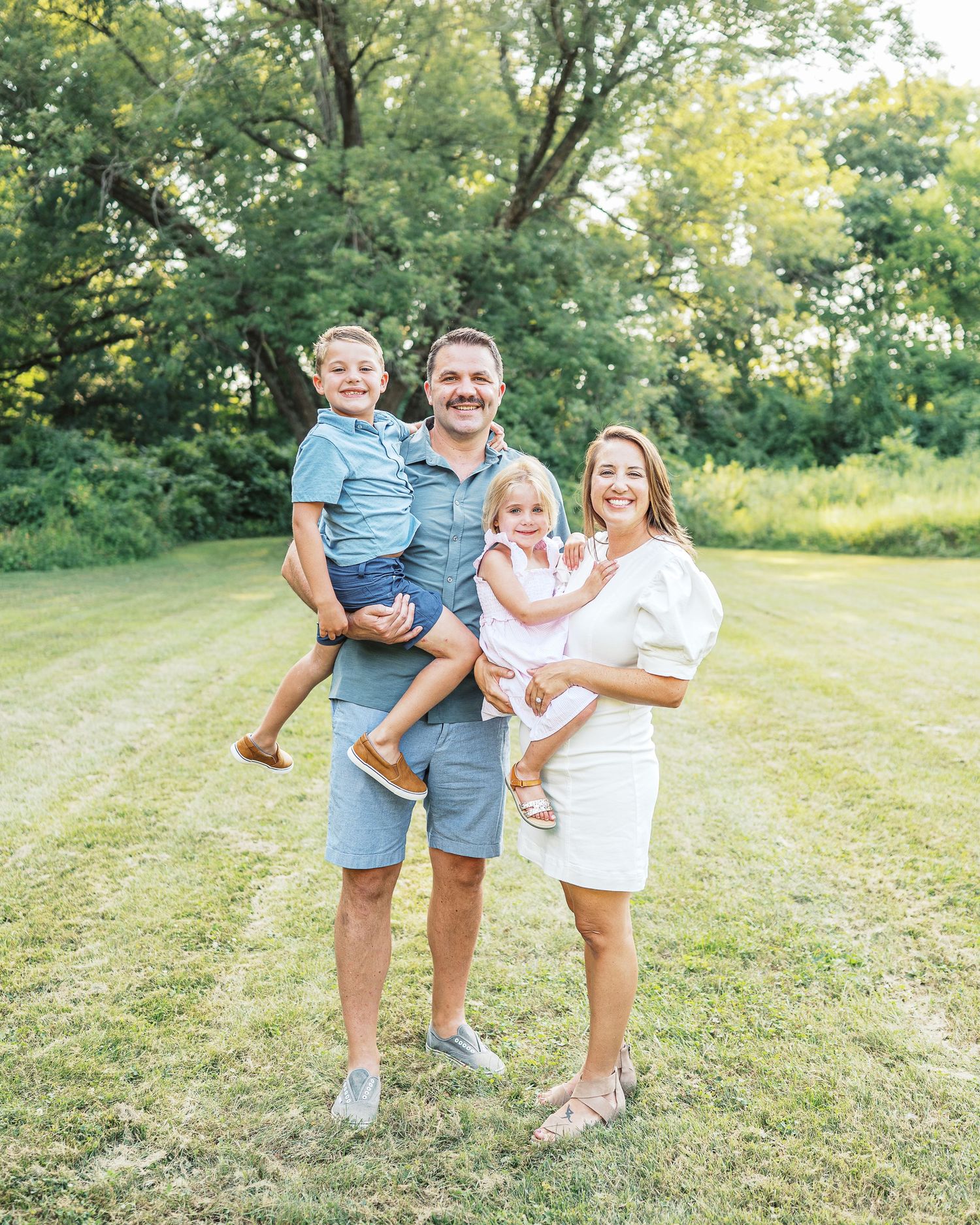 Happy family posing together in a sunny meadow with green trees in background.