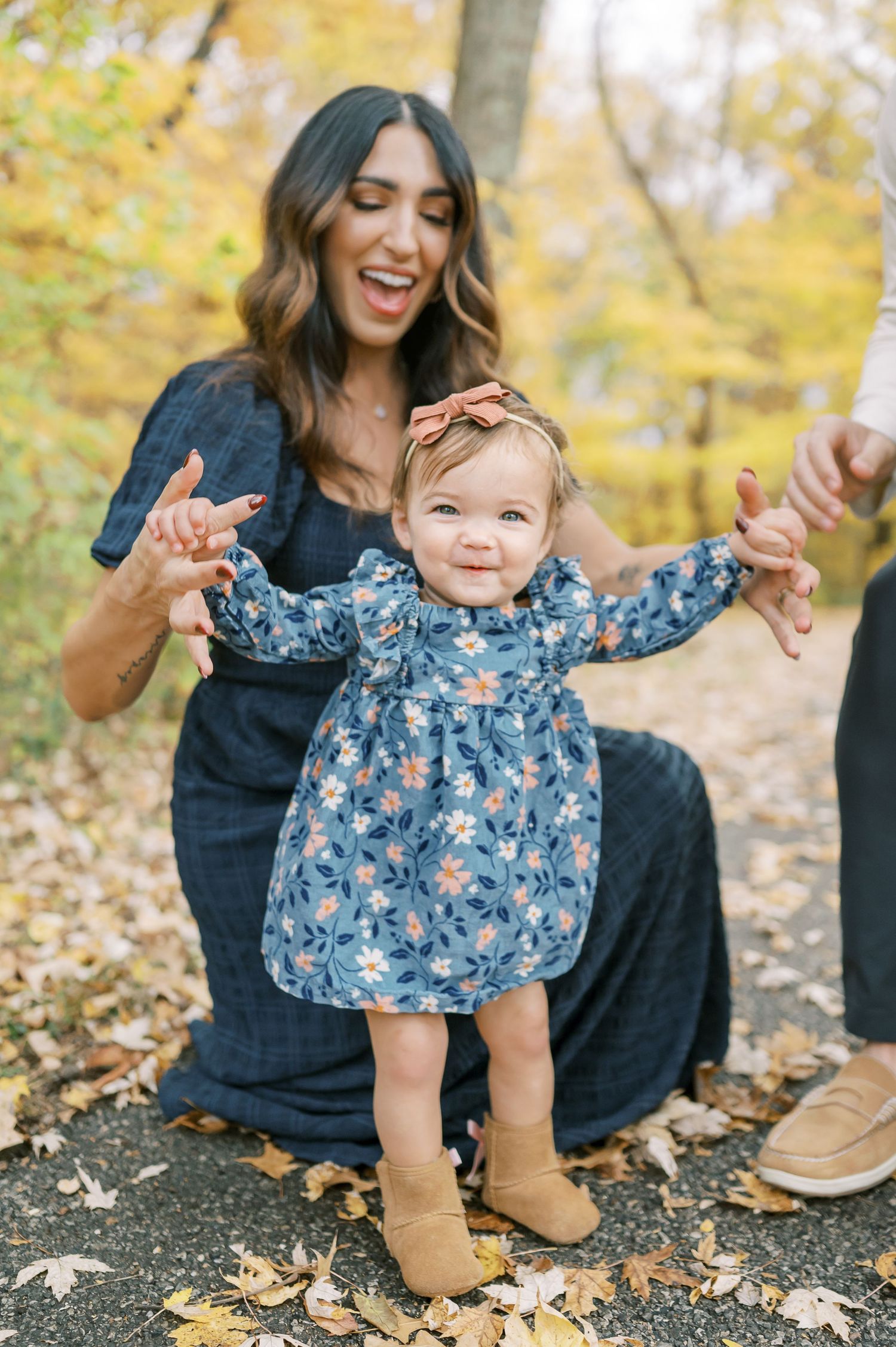 Young child in blue floral dress learning to walk while holding hands with parent outdoors.
