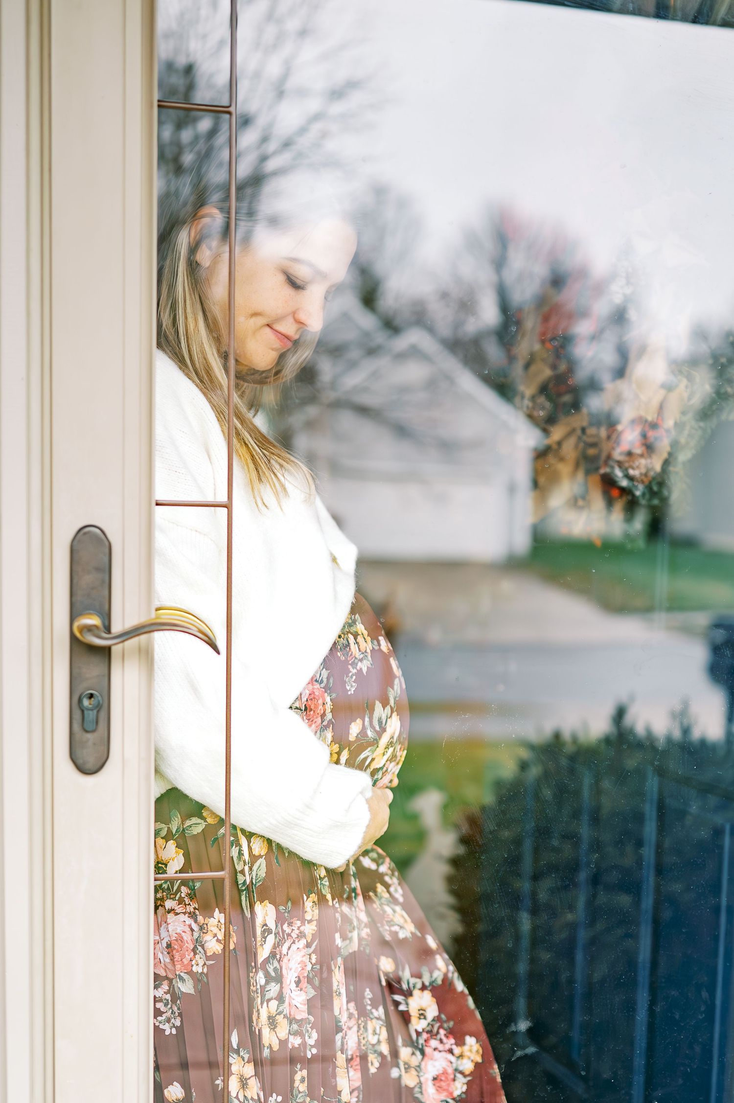 Side profile of expectant mother in floral dress standing by white door in natural light.
