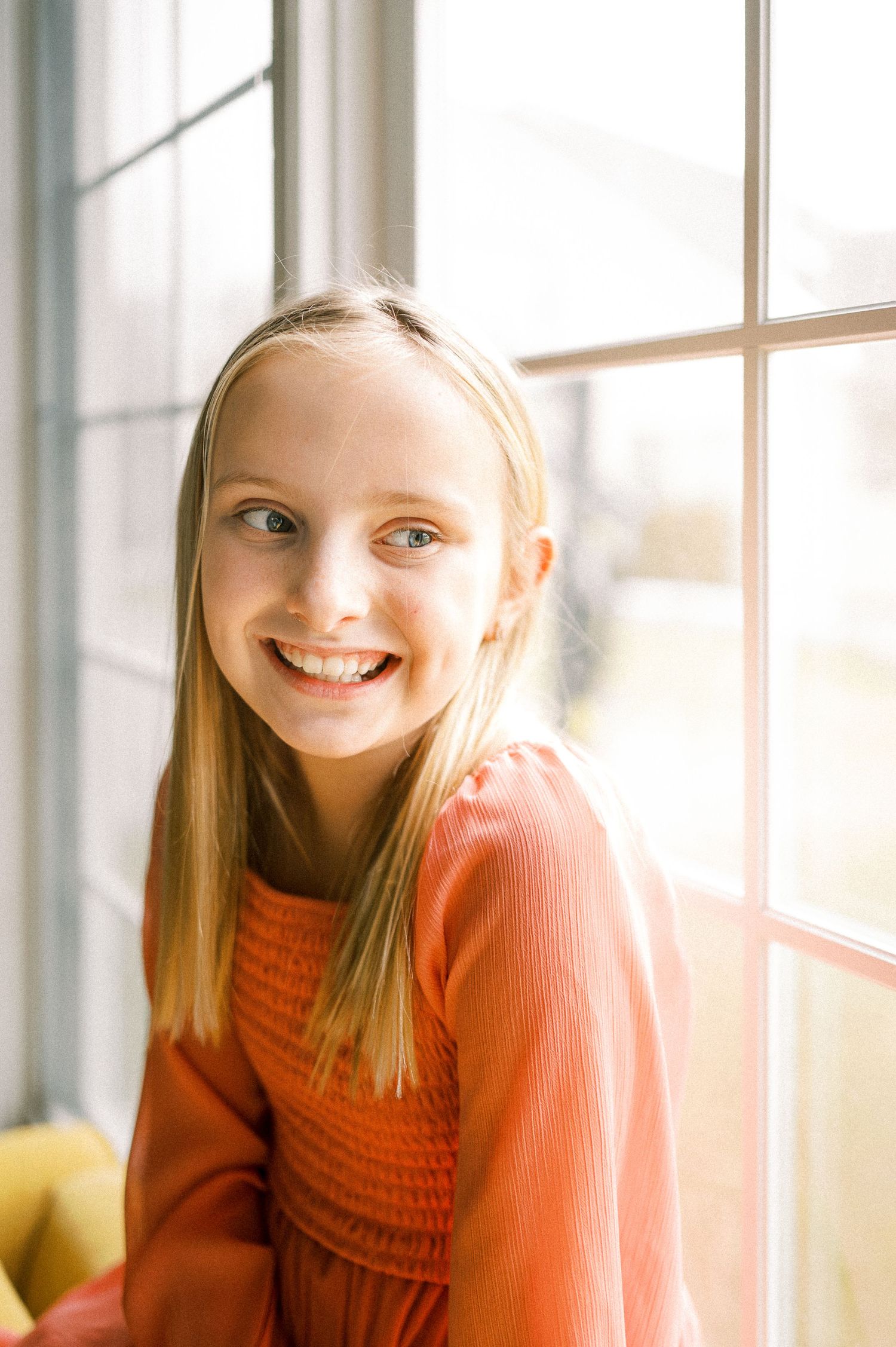 A person in a coral sweater smiles warmly by sunlit windows.