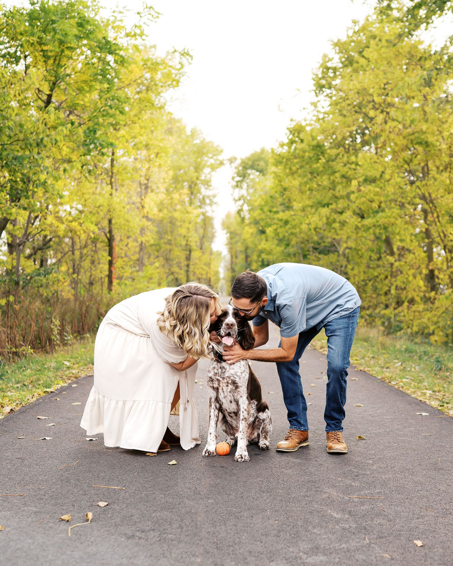 Two people interact with their spotted dog on a paved path surrounded by fall foliage.