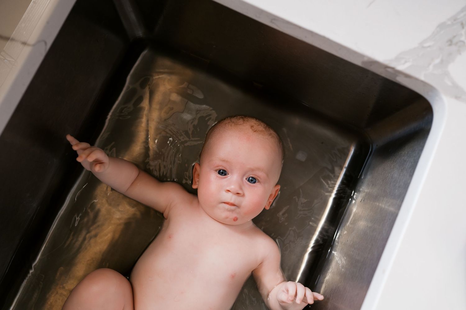 Baby resting in stainless steel kitchen sink during bath time.