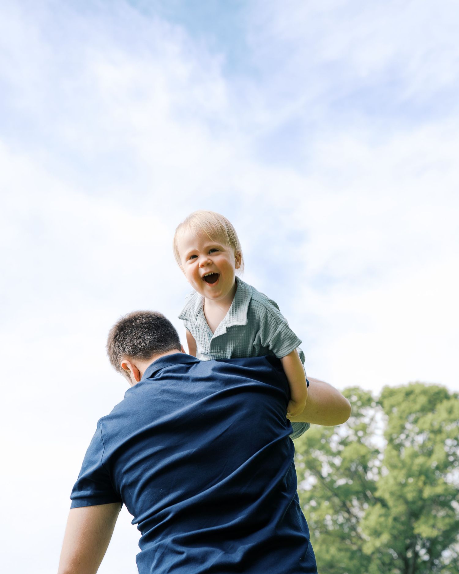 Toddler in blue shirt laughing joyfully while being lifted up in sunny outdoor setting.