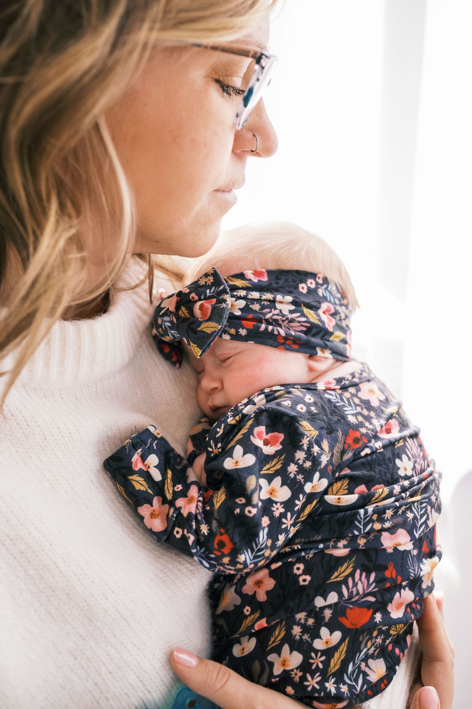 Mother in glasses holding sleeping baby wrapped in floral print blanket.