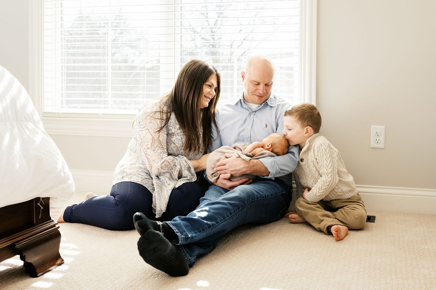 A family sits together on carpet floor while bonding with their newborn baby in a bright room.