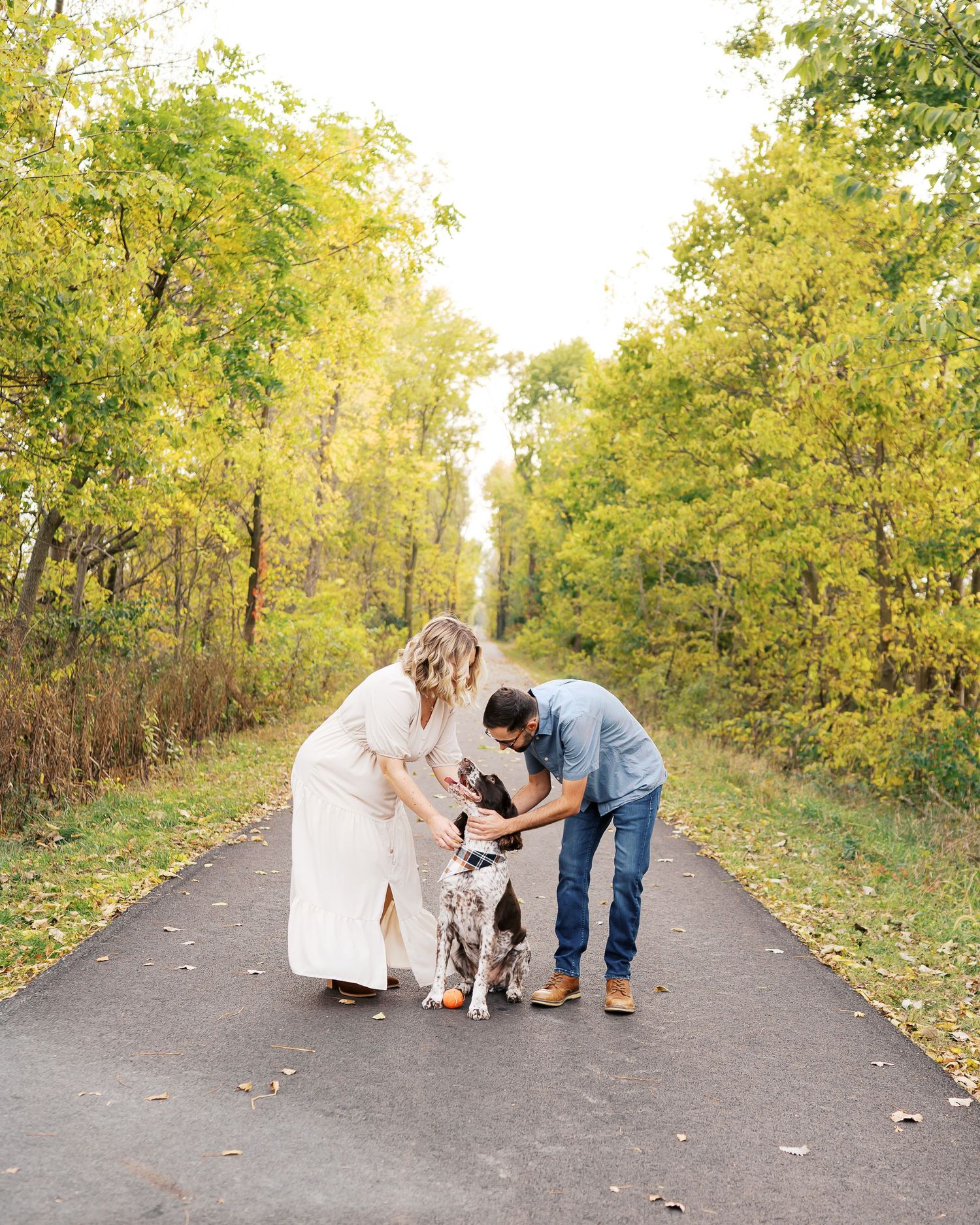 A couple kneels down to pet their dog on a tree-lined path covered in fallen autumn leaves.