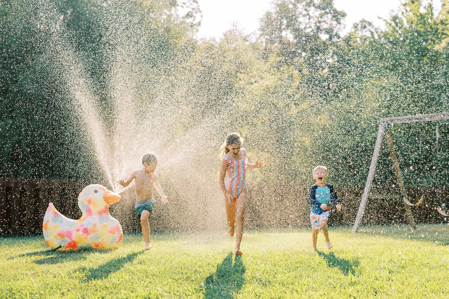 Kids running through water sprinklers with an inflatable pool float in a sunny backyard.